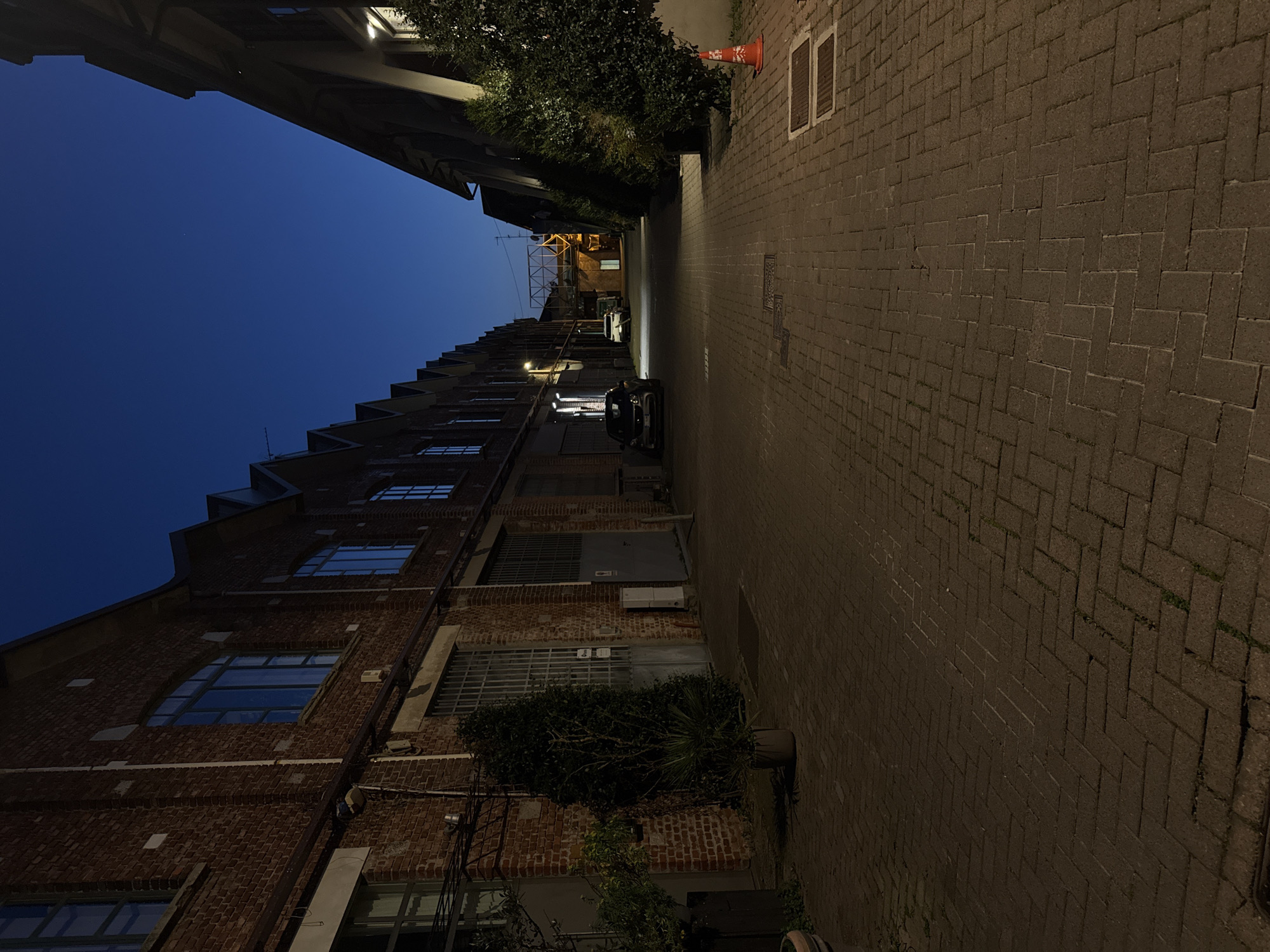 The image depicts a narrow street in Milan, Italy, at twilight. On one side, a tall brick building with multiple windows rises, some covered with metal grates. The architecture shows a stepped roofline. On the opposite side of the street, a lighter-colored building is visible. The street is paved with herringbone-patterned bricks. An orange traffic cone is placed at the edge of the sidewalk. Several vehicles are parked further down the street. The sky is a deep blue. Some greenery is visible along the buildings.