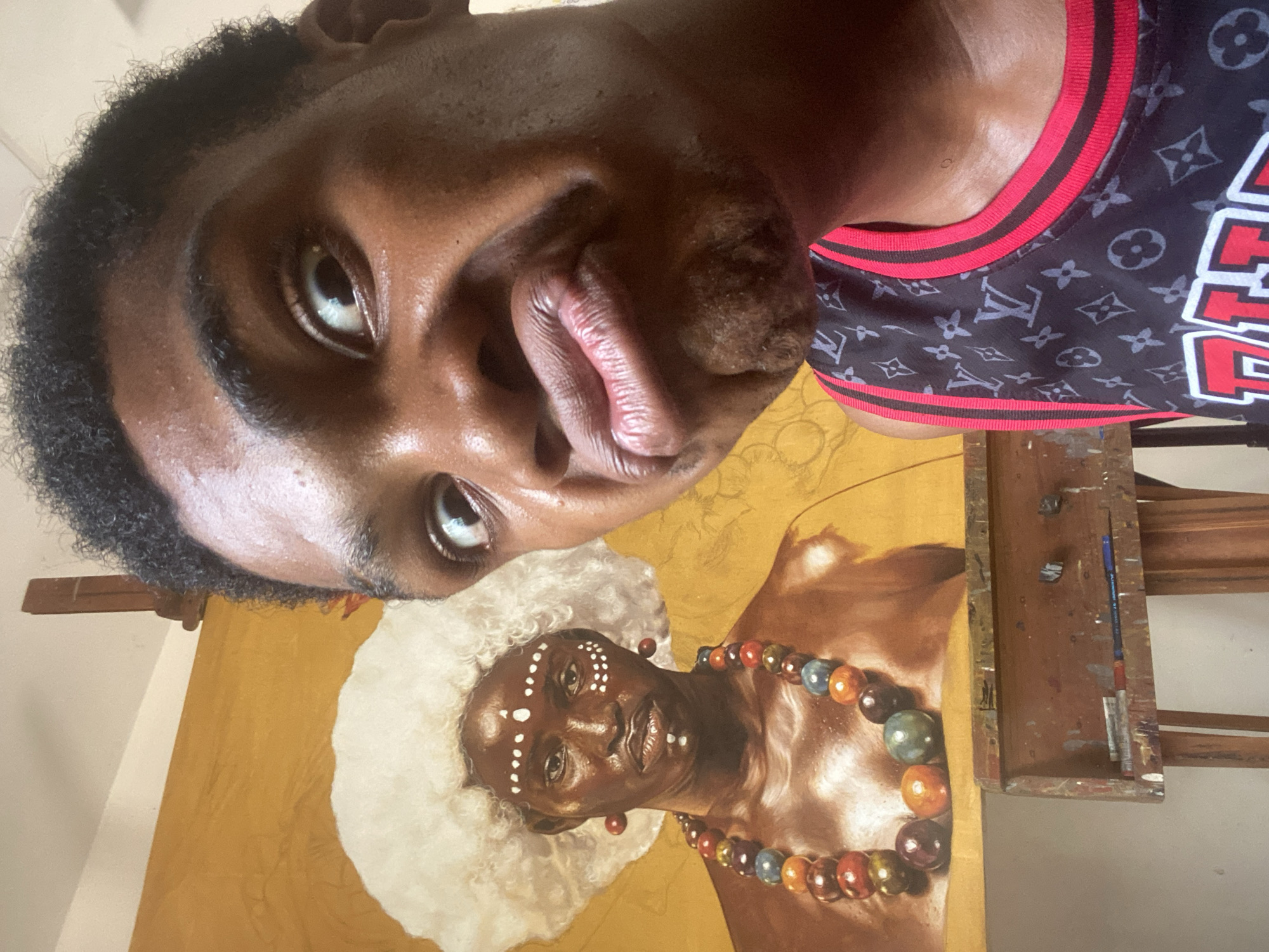 A selfie photograph, taken from a low, upward angle, captures a young Black man looking directly at the camera, his face filling the upper portion of the frame. He has short, curly dark hair and a thoughtful or slightly playful expression with pursed lips. He wears a black basketball-style jersey with red and white trim, featuring Louis Vuitton monograms and partially visible red text on the shoulder that appears to read "ALLE."
Below the man, a large, detailed painting is displayed on a wooden easel. The artwork is a portrait of an adult Black individual with dark skin, adorned with distinctive white facial markings (dots and lines) and a white, textured hairstyle resembling an afro. The painted subject wears a prominent, multicolored beaded necklace with large, round beads in various hues, along with red disc earrings. The background of the painting is a solid golden-yellow.
The setting is an indoor space, likely an artist's studio in Ado, Nigeria, given the provided context. On the lower shelf of the easel, paint tubes, a pen, and brushes are visible, indicating an active workspace. The lighting suggests it is daytime. The image captures the artist presenting himself alongside his culturally rich and visually striking artwork.