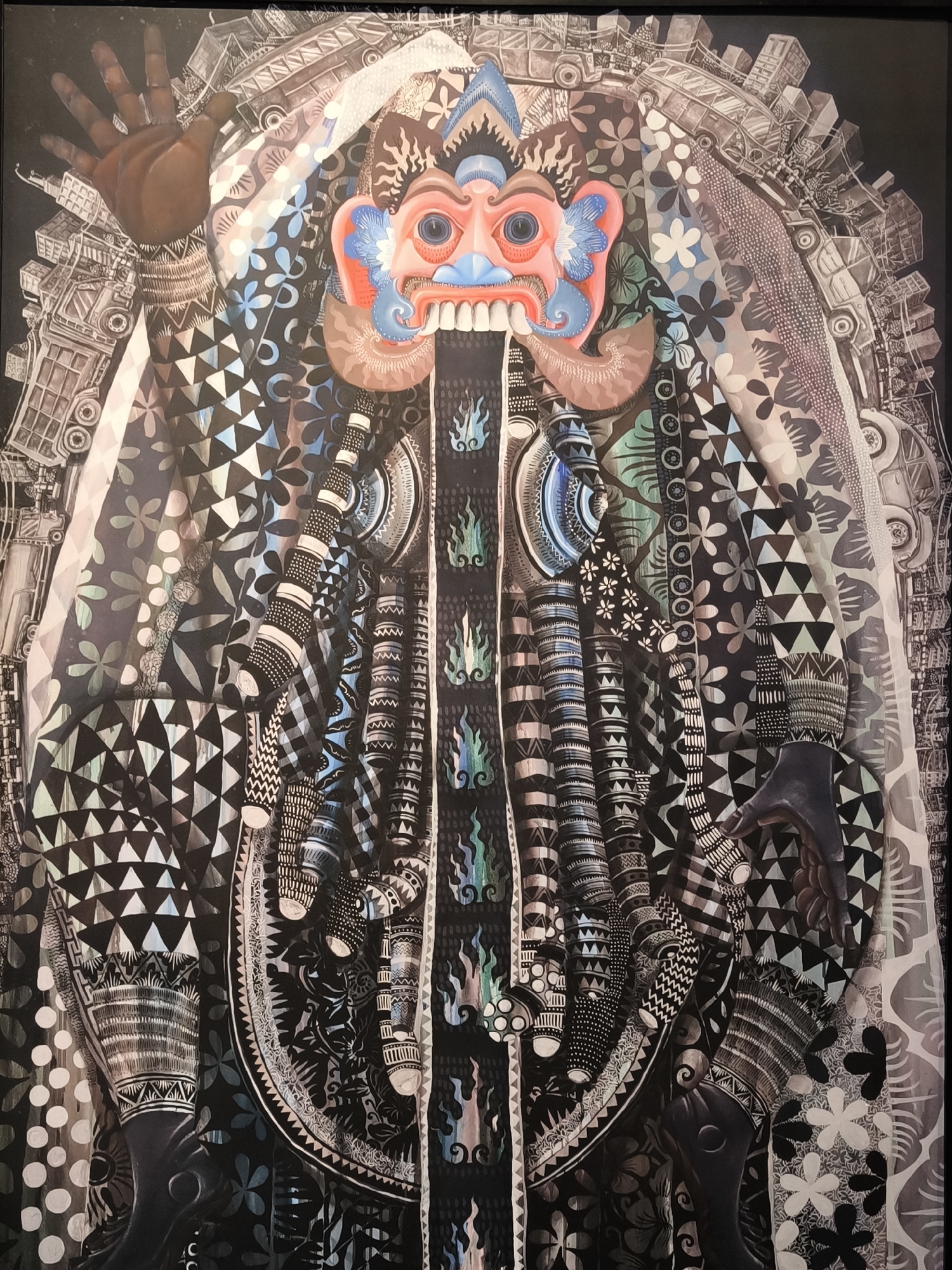 A detailed, colorful artwork shows a complex, abstract figure with a prominent mask-like face at the top. The face has a pink complexion, blue accents, large eyes, and a wide, toothed grin. Below the face, a long, dark panel descends, adorned with flame-like motifs in blue and green.
The figure is draped in layers of patterned fabrics. Geometric patterns, floral designs, and stripes are incorporated into the textiles. On either side, hands reach into the frame, with one raised in a gesture. An overlaying background suggests a cityscape composed of vehicles. The overall color palette combines dark and light tones, with pops of bright color concentrated around the mask.
Given the art style, location of Mekarsari, and possible Indonesian cultural connection, it potentially draws on local folklore and religious themes, although this can't be stated definitively without additional context.