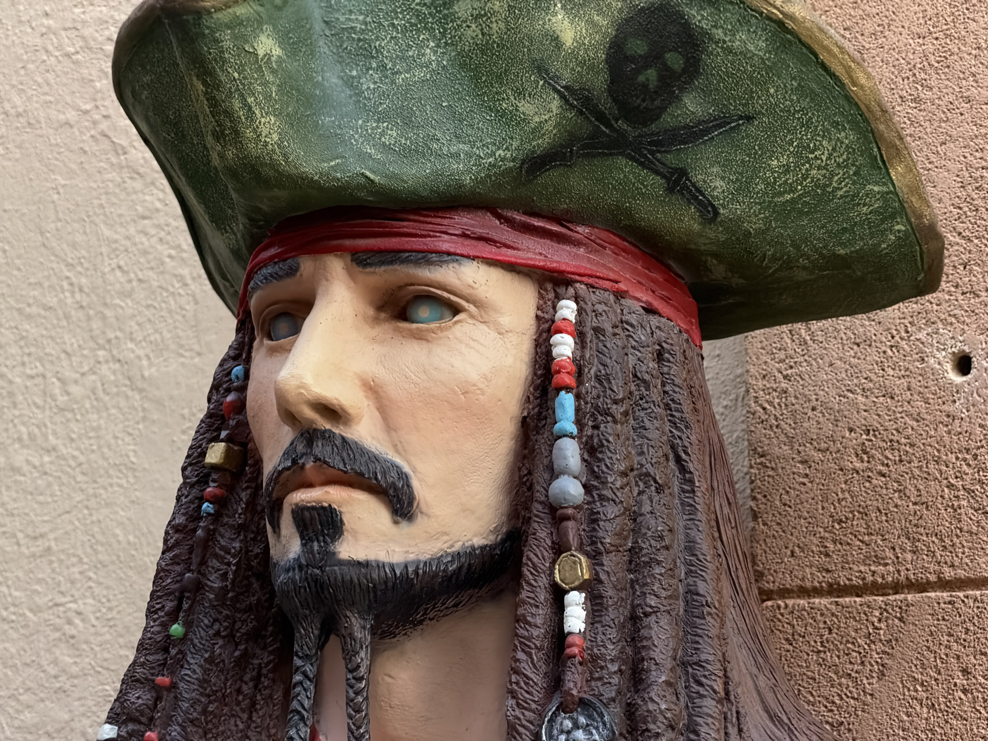 A close-up shot of a Jack Sparrow bust outdoors. The bust has a green pirate hat with a skull and crossbones, a red bandana, and a brown beard with beaded braids. The bust appears to be made of plaster or a similar material and is set against a neutral wall. The location is Alcúdia, Spain.