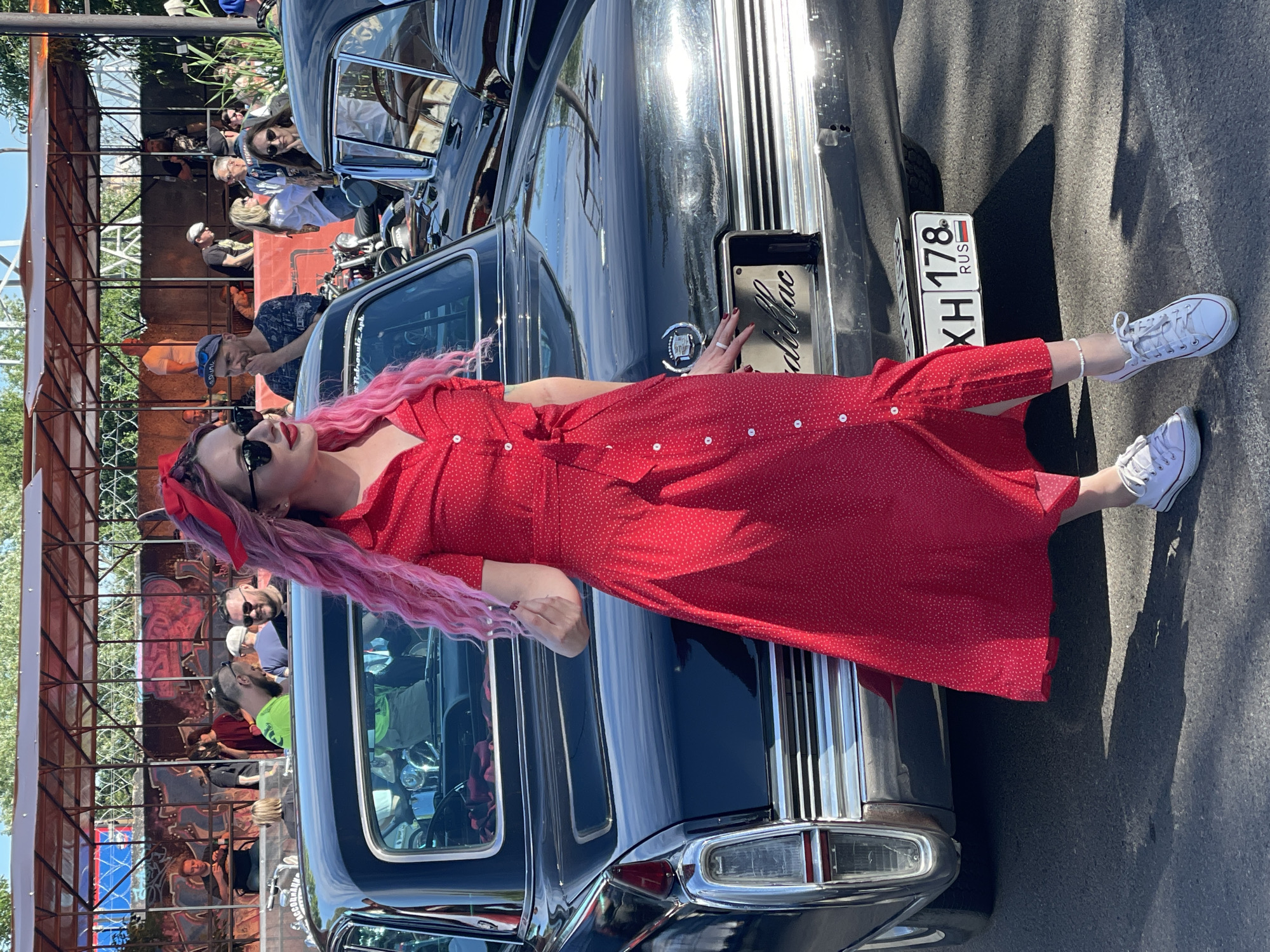 A woman with bright pink hair and a red polka dot dress stands next to a black vintage car. She wears sunglasses and a red headscarf. The car has a license plate that reads "XH 178 RUS". In the background, other people and cars are visible, as well as a covered structure with graffiti on the wall behind it. The scene appears to be outdoors in a city setting, possibly at a car show or event. The license plate's format suggests the location is indeed Saint Petersburg, Russia. The overall atmosphere is casual and summery.