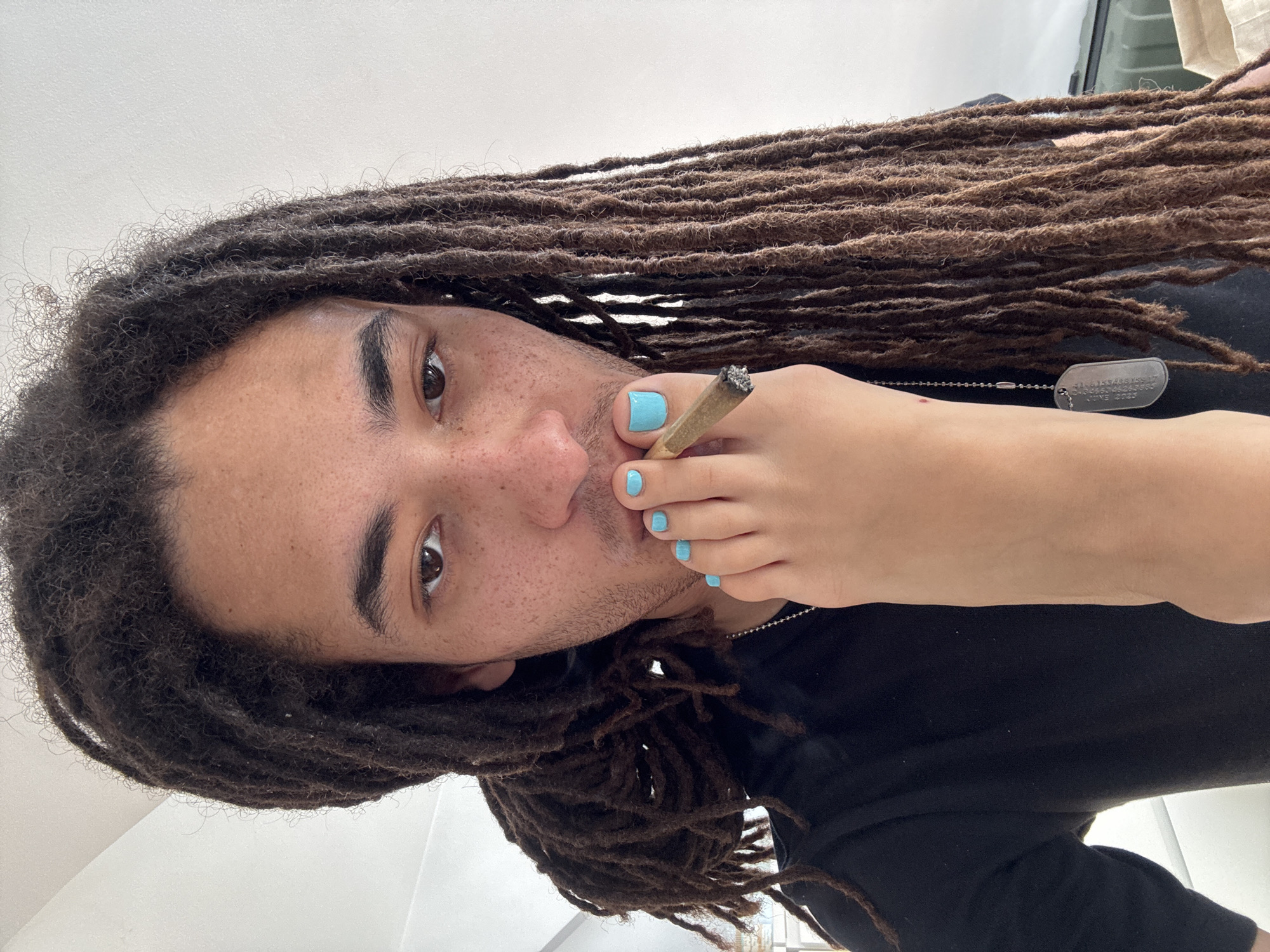 A close-up, angled shot features a person with brown dreadlocks and light brown skin, holding a foot with polished light blue toenails up to their mouth. The person is holding a lit cigarette between the big toe and second toe of the foot. The person is wearing a black shirt and a metal dog tag necklace. The background is blurred and features a window. The location is not discernible as Paris, France, from the visual information provided.