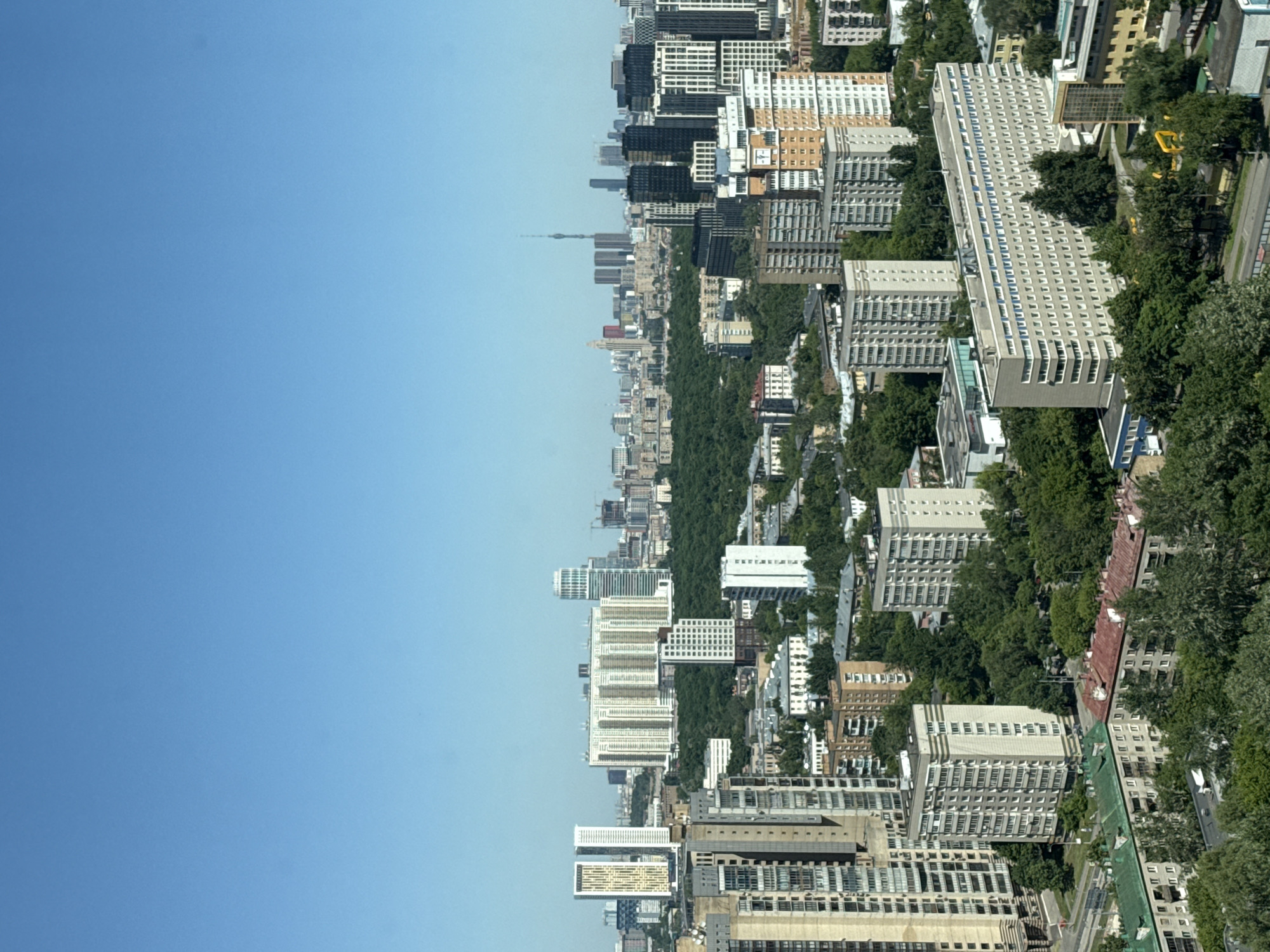This image presents a high-angle, wide-view shot of an urban landscape under a clear, cloudless blue sky. The sky, occupying the top third of the frame, transitions from a deeper blue at the zenith to a lighter hue near the horizon.
In the far distance, a discernible city skyline is visible, characterized by numerous tall buildings and skyscrapers. A prominent, slender telecommunications tower, consistent with the Ostankino Tower, is identifiable slightly right of the frame's center on the horizon line. This distant cluster of high-rises strongly suggests the city of Moscow.
The mid-ground is dominated by a substantial, dense expanse of dark green trees, likely a large park or forest. Directly in front of this green belt, several large, modern residential complexes stand out. On the left, two tall, light-colored buildings with distinct vertical patterns are visible. To their right, a sprawling complex of light-toned, multi-story residential buildings with numerous windows forms a prominent structure, adjacent to a taller, cylindrical white skyscraper with a rounded top.
The foreground and closer mid-ground feature a mix of residential buildings. On the left, a tall, beige-colored apartment building with many windows is present. In the center-right foreground, a long, rectangular, multi-story building with a light gray facade and a large number of uniformly spaced windows is prominent. Scattered throughout are numerous older, block-style residential buildings, typically 5 to 12 stories high, with flat or gently sloped roofs, some green and some reddish-brown, nestled among abundant mature trees. Roads are partially visible winding between buildings and through green spaces.
No individuals or vehicles are clearly discernible due to the elevated vantage point. The scene is brightly illuminated by natural sunlight, indicating clear weather during late morning or early afternoon. The combined architectural styles, the extensive greenery, and the identifiable distant Moscow skyline, including the Ostankino Tower, place this scene within the city of Khimki, Russia, which borders Moscow to the northwest.