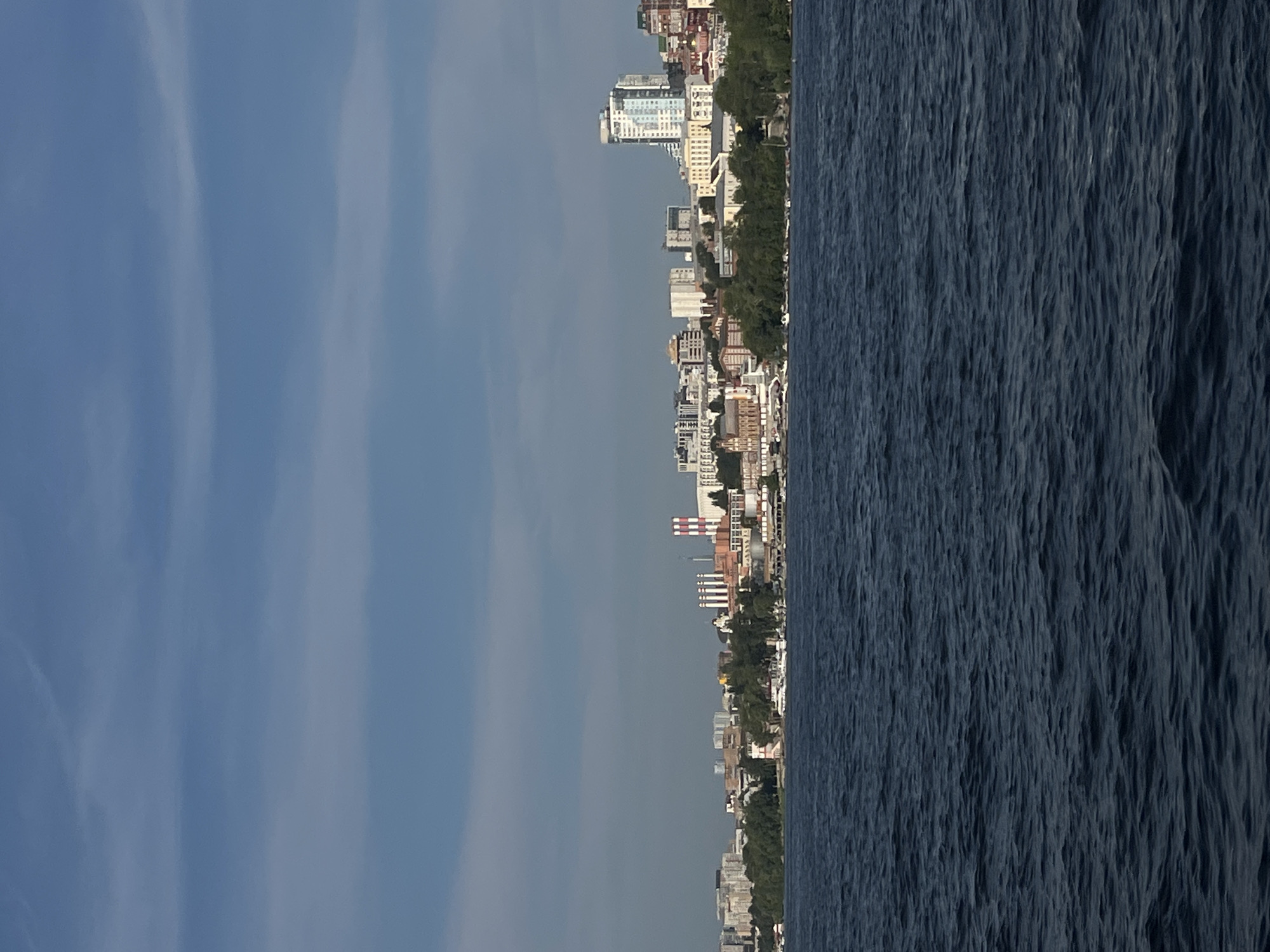 The image shows a panoramic view of the Novokuybyshevsk skyline from the water. The foreground is dominated by dark, choppy water. In the middle ground, a city stretches along the shoreline, characterized by a mix of buildings, including residential towers, industrial structures with tall chimneys, and older, lower-rise buildings. Lush green trees line the waterfront, interspersed with the urban landscape. The sky above is a clear blue with faint, wispy clouds. Several boats are visible docked along the shore. The overall impression is that of a coastal city with a blend of modern and older architecture.