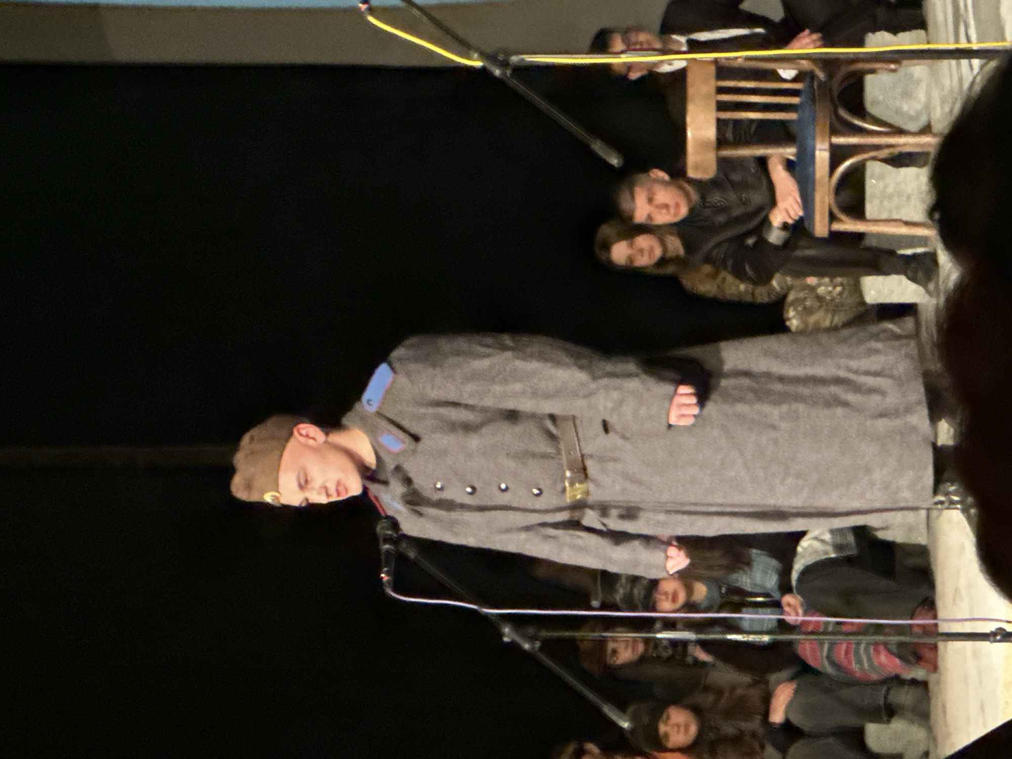 In Panagyurishte, Bulgaria, a male dressed in a military uniform is positioned on stage in front of a microphone. An audience is present, with people visible in the foreground and background. A chair is visible in the background. The setting appears to be a performance or presentation.