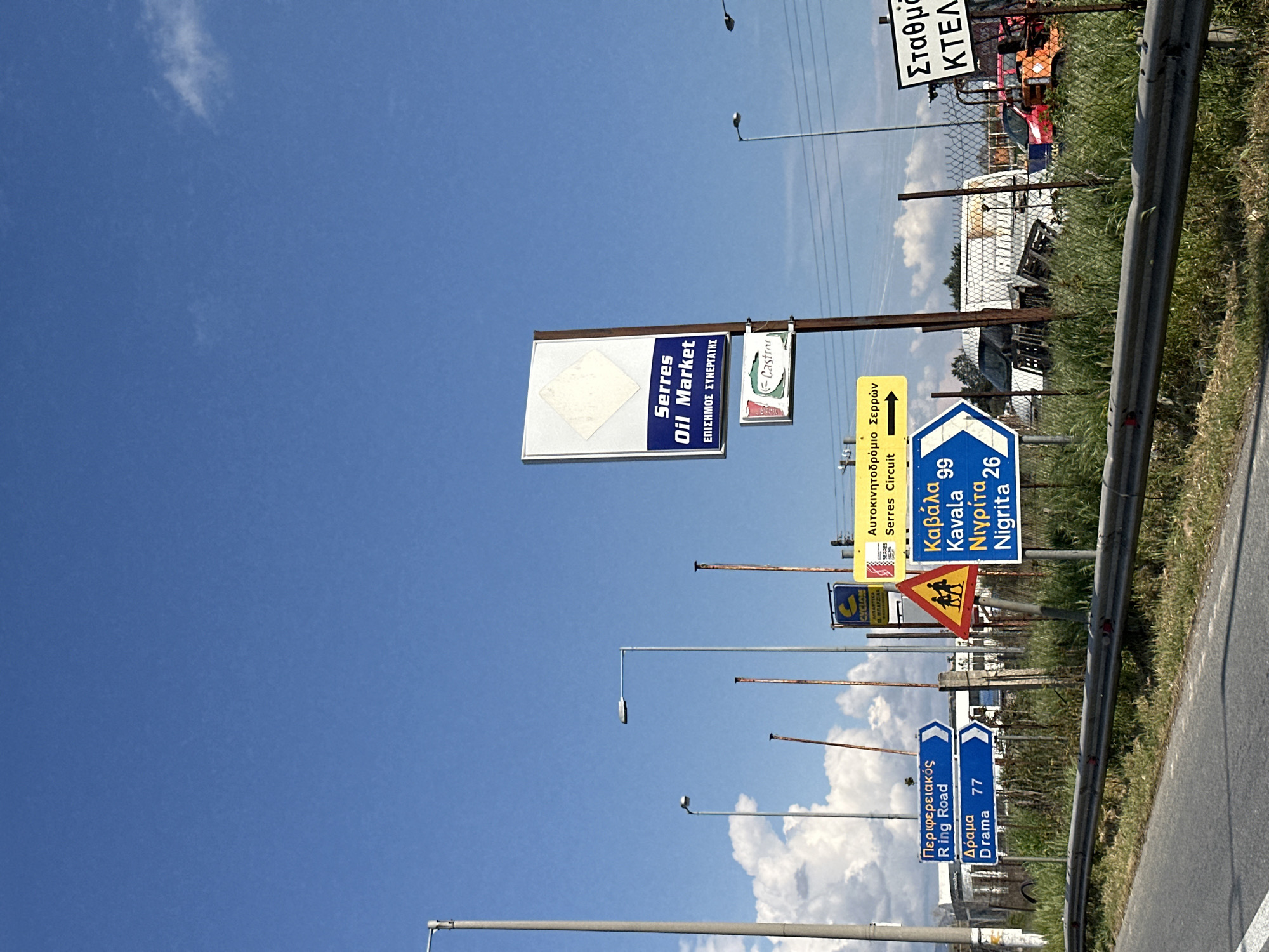 The image shows a series of roadside signs under a bright blue sky in Lefkonas, Greece. The signs include directions and advertisements. There is a sign for "Serres Oil Market", followed by other signs indicating directions to "Serres Circuit", "Kavala", "Nigrita", and "Drama". One sign reads "Ring Road" and "Drama 77." There is also a sign showing a school crossing and another one for "Ktel Station". These signs are arranged along a highway near a metal barrier, indicating this is likely a roadside scene on the outskirts of the city.