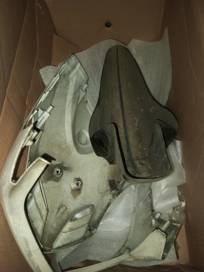 A cardboard box contains motorcycle fairings. A larger, off-white piece displays visible attachment points and a front-facing design. A smaller, dark gray piece rests atop it. The pieces are surrounded by gray packing material. The box is brown with a dark circular mark in the upper corner. The fairings appear used, with visible dirt and smudges on their surfaces.