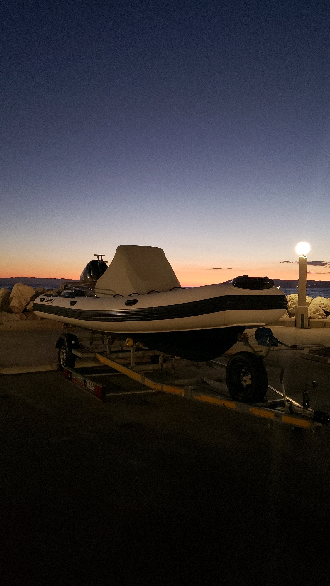 An inflatable boat on a trailer is parked on a paved surface near the sea at dusk. The boat is white with a black stripe and covered with a tan tarp. The trailer is yellow metal with black tires and yellow reflective tape. In the background, large rocks line the shore, and the sea is visible. A lamppost with a lit bulb stands on the right side of the frame, casting light on the scene. The sky transitions from a deep blue at the top to orange and pink hues near the horizon, indicating sunset or sunrise. The location is identified as Općina Tučepi, Croatia. There are no people visible in the image.