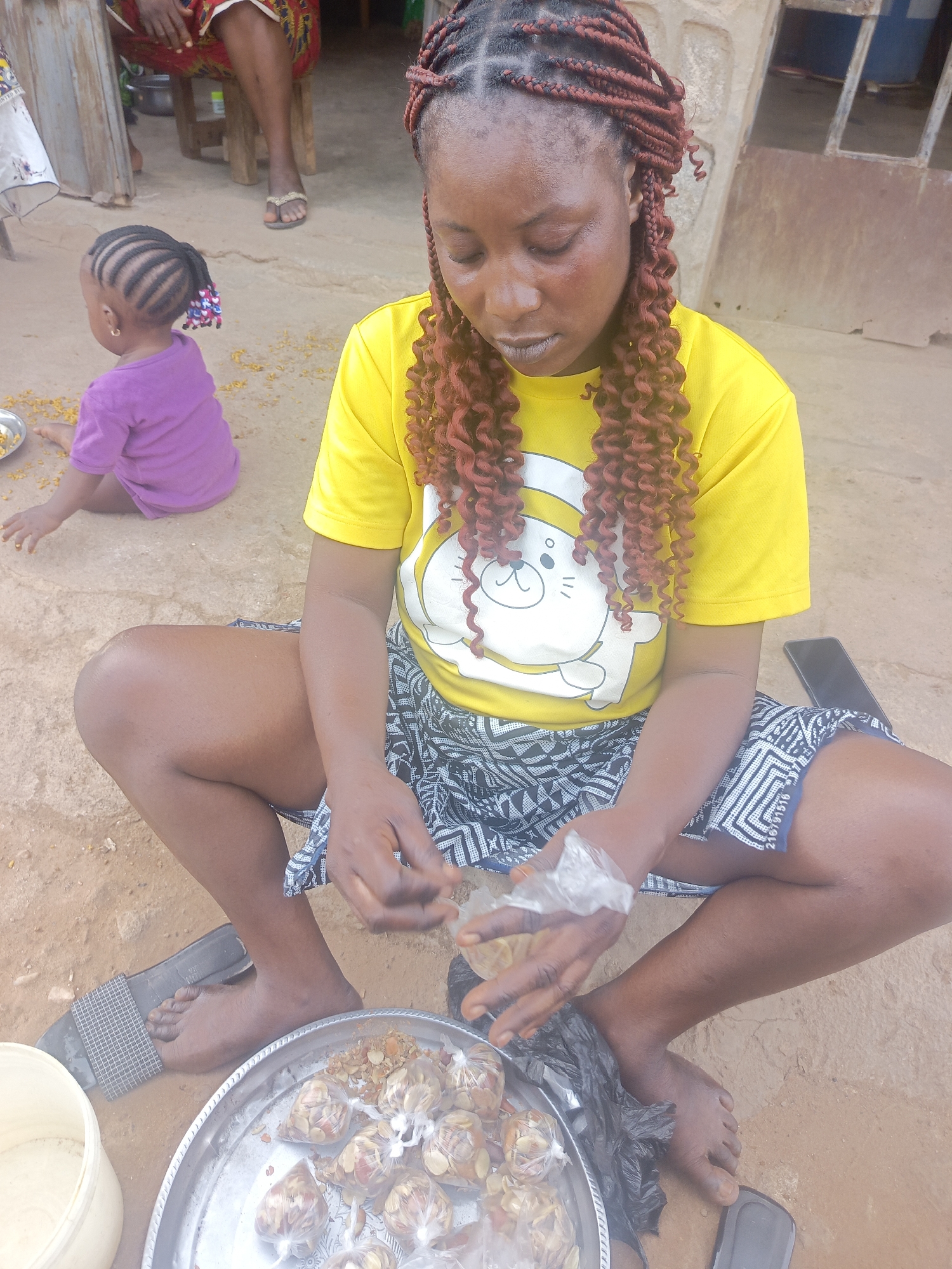 A woman with reddish-brown braided hair wearing a yellow t-shirt with a cartoon graphic and a black and white patterned skirt is sitting cross-legged on the ground. She is packing food items, possibly peanuts, into small plastic bags. A silver tray filled with pre-packaged bags of the same food item is near her. To her left is a white plastic container.
A young child with braided hair and a purple t-shirt is sitting in the background.
The ground is a dusty brown color, suggesting an outdoor setting. A black sandal is visible near the woman's foot.
The presence of the child and the food items suggests a domestic or market-related activity. It's impossible to pinpoint the exact location within the city of Akwanga, Nigeria without additional information.