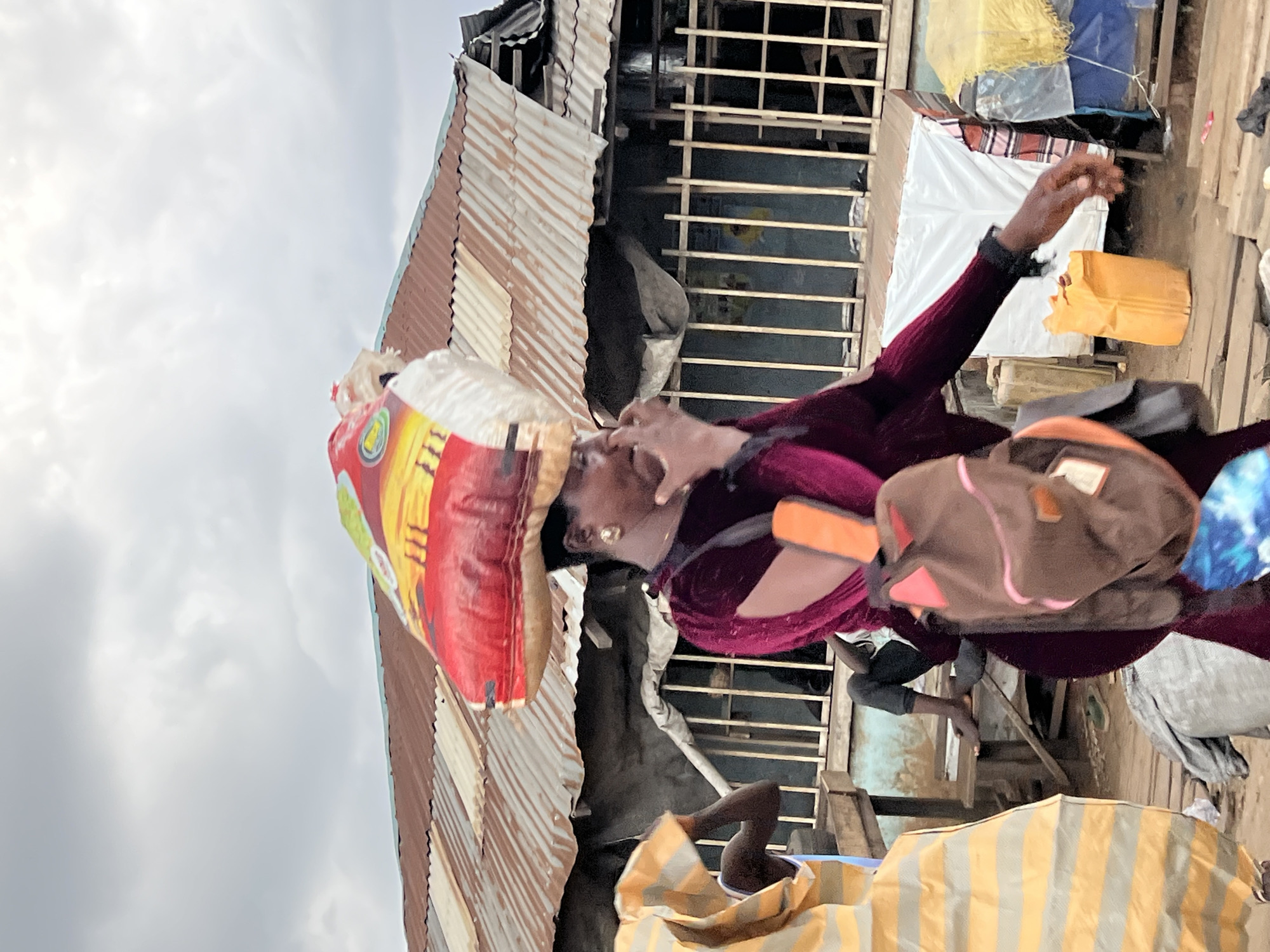 In Oko Amakom, Nigeria, a woman with dark skin wears a dark maroon velvet dress with cutouts on the sleeves. She carries a large bag of rice on her head and a brown backpack with pink accents over her shoulder. Her right hand is near her face, possibly touching her nose or adjusting her mask. In the background, a dilapidated building with a rusted corrugated metal roof is visible. A man sits on a wooden structure in front of the building, his legs bare. Another person, mostly obscured, wears a yellow and orange striped garment on the left side of the frame. The ground is made of wooden planks, and various objects, including an orange plastic container and a grey tarp, are scattered around. The sky is overcast with clouds.