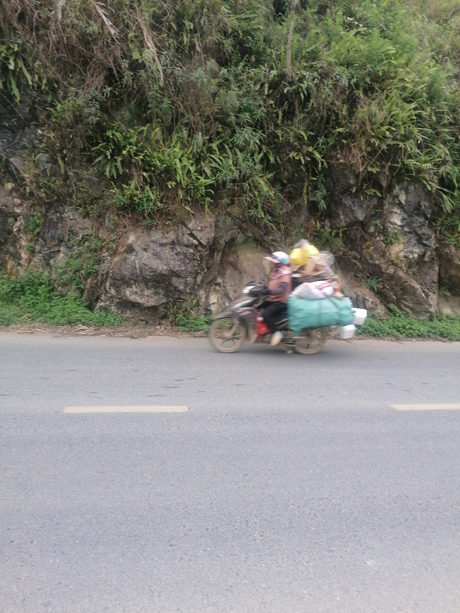 The image captures a moment on a paved road, likely in a rural or mountainous area such as Sa Pa Ward, Vietnam. The central subject is a motorcycle in motion, evidenced by the significant motion blur affecting the vehicle and its occupants.
One person, presumably the driver, is visible on the motorcycle, wearing a light-colored helmet or cap and a reddish-brown or pinkish long-sleeved top. The motorcycle itself is heavily laden with cargo, characteristic of transportation methods in the region. A large, dark green tarpaulin-wrapped bundle is secured to the rear of the bike, extending considerably beyond its frame. Above this, several other items are stacked, including a prominent yellow bundle and lighter-colored bags, suggesting a substantial amount of goods being transported.
The road is an asphalt surface, grey in color, with a single faded yellow dashed line running horizontally across its lower half. To the left of the road, a steep, rocky embankment rises, densely covered with varied green foliage, including ferns and shrubs, interspersed with bare rocks and dried branches. The vibrant green of the plants contrasts with the darker grey of the rocks and the road.
The scene depicts a common sight of practical transport, where motorcycles are utilized for carrying heavy loads through what appears to be a natural and rugged landscape. The lighting suggests it is daytime, possibly under an overcast sky, as there are no distinct shadows. No visible text is present in the image.