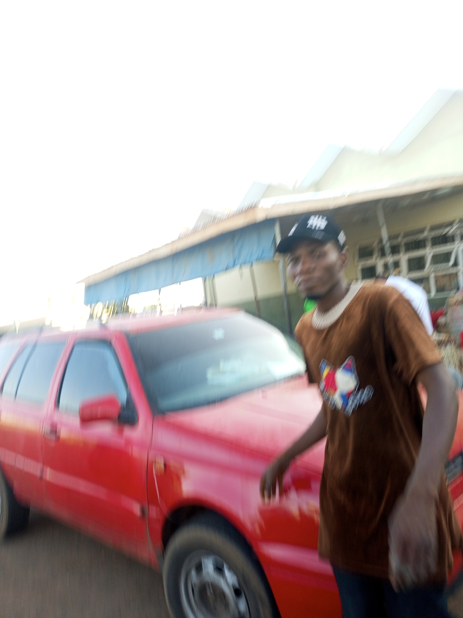 A blurry photograph depicts a man standing beside a red car. The man is wearing a brown t-shirt with a graphic design and a black baseball cap. He is positioned on the right side of the car, appearing to lean on it. The car is a red station wagon, with the front left side visible. In the background, there is a building with white walls and windows, and a blue awning. The location is in the city of Unknown, Unknown.