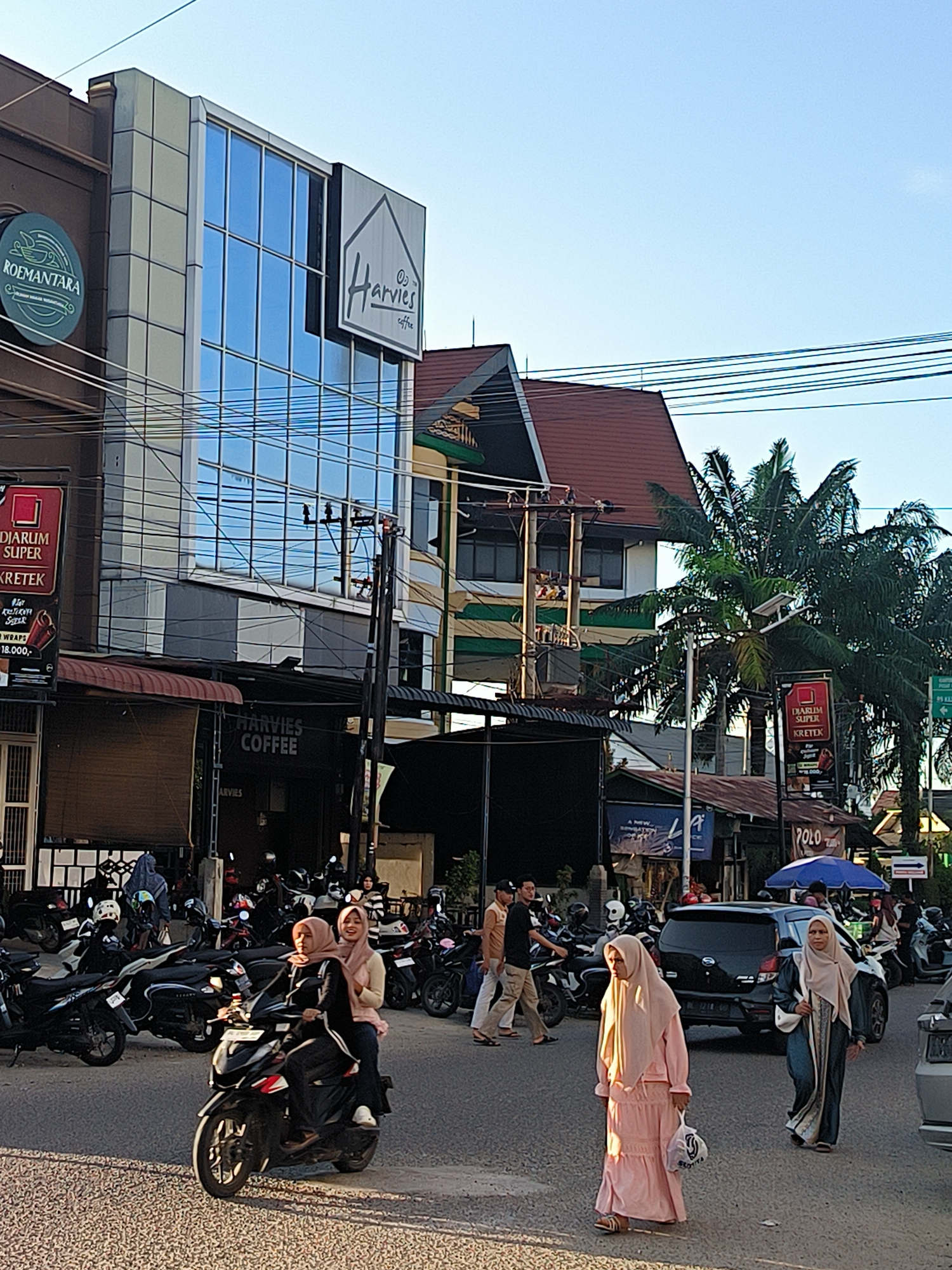 The image captures a street scene in Lhokseumawe, Indonesia. The foreground shows a crosswalk where two women, dressed in traditional Muslim attire, are walking. One woman is wearing a pink dress with a beige hijab, and the other a blue and beige outfit with a beige hijab. To the left, two other women are riding a scooter. The driver wears black clothing and a beige hijab, while the passenger wears a pink shirt and a beige hijab.
In the background, numerous parked motorcycles line the street on both sides. Several pedestrians are also visible. Shops and buildings form the backdrop, including "Harvies Coffee", which appears as both a storefront and on a sign on a taller building above, and the words "ROEMANTARA" in a circle on another building. A palm tree is visible to the right, and there are also signs advertising "DJARUM SUPER KRETEK" and "POLO". Overhead, power lines crisscross the sky.