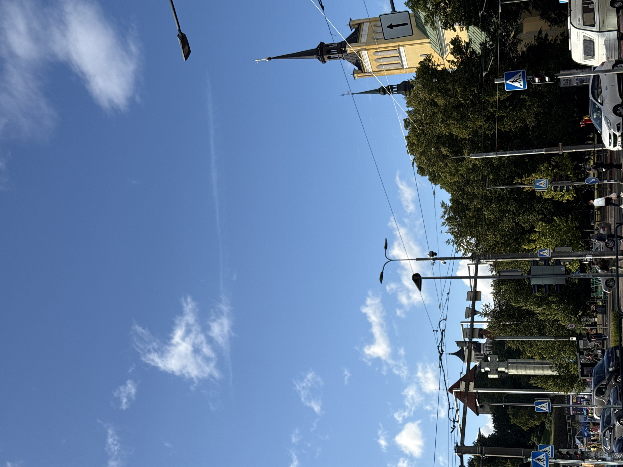 The image depicts a street scene in Tallinn, Estonia, with a clear blue sky dotted with white clouds. In the foreground, a street intersection is visible with traffic lights, pedestrian crossings, and several vehicles, including cars and a white caravan. Traffic signs indicating pedestrian crossings and directions are also present. Overhead, a complex network of trolleybus wires spans the scene.
Dominating the middle ground and background are lush green trees, behind which several notable structures are partially visible. On the right, a yellow church with a tall black steeple topped with a cross stands out. Further to the left, glimpses of older, stone buildings with red roofs suggest historical architecture, possibly part of Tallinn's Old Town. A tall, cylindrical monument with what appears to be a cross or monument on top is also visible amongst the trees.
Several individuals are present, walking on the sidewalks and crossing the street. They appear to be engaged in everyday activities such as commuting or sightseeing. The overall impression is of a typical urban environment in a European city, with a blend of modern infrastructure and historical landmarks.