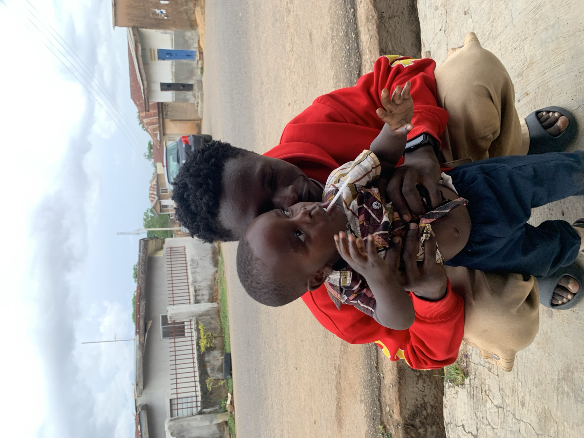 A man and a child are positioned on the side of a road in a residential area. The man, wearing a red sweatshirt, is holding the child, who is wearing a plaid shirt and dark pants. The man is also wearing dark-colored sandals. In the background, there are buildings with various architectural styles and a gray vehicle parked along the side of the road. The road appears to be paved and has some discoloration or staining. The sky is overcast. The location is Jos, Nigeria, but further details are unavailable.