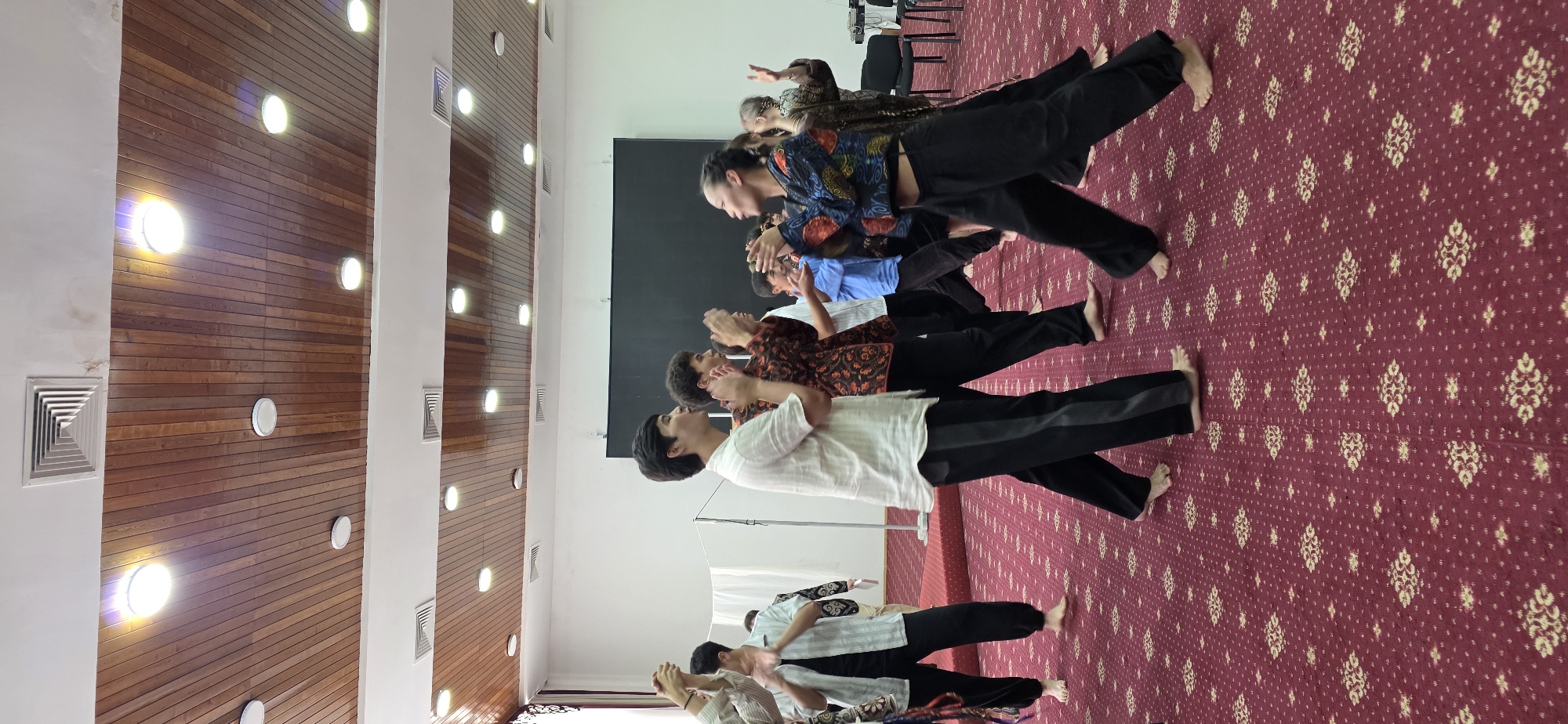 The image shows a group of people clapping their hands in an indoor space with a red patterned carpet and a wooden ceiling with circular lights. A few people are in focus in the foreground, including a man in a white shirt and black pants, and a woman in a colorful patterned top and black pants. They appear to be participating in a dance or performance. In the background, other individuals are also clapping. The room has white walls and a large black screen at the back. The overall scene suggests a cultural event or rehearsal in Bulan-Segettu, Kyrgyzstan.