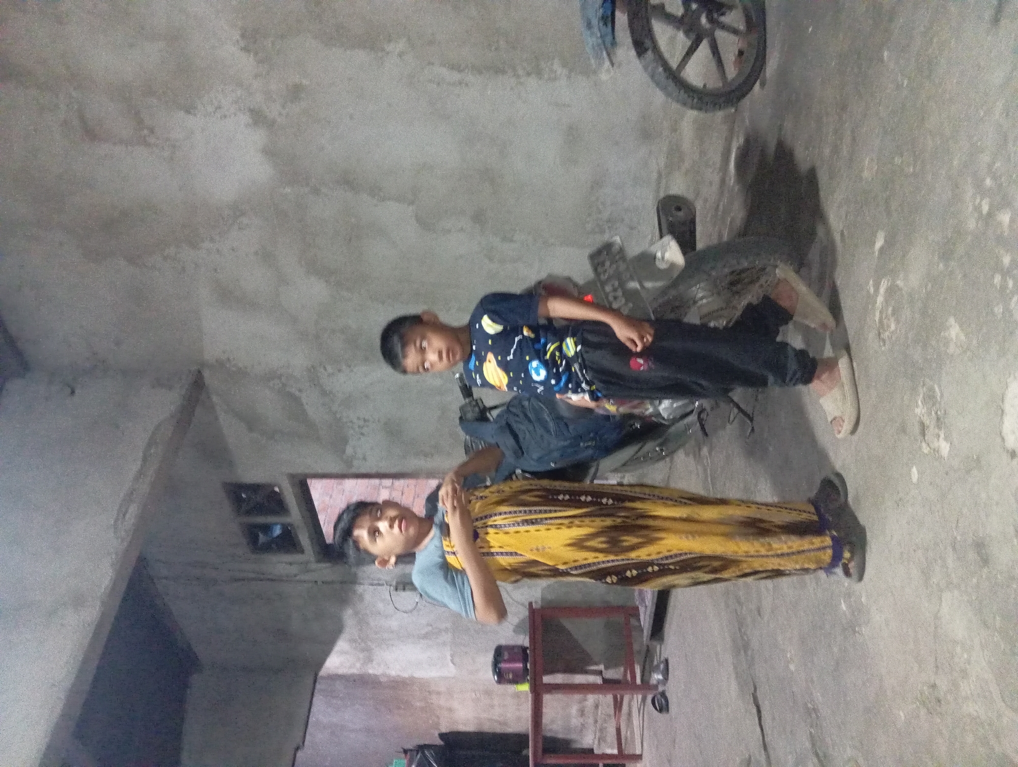 Two young boys are standing in what appears to be a dimly lit, unfinished building or outdoor covered area. The boy on the left is wearing a light blue t-shirt and a long, patterned yellow and brown sarong. He is holding his hands crossed in front of his chest. The boy on the right is wearing a dark blue t-shirt with a space-themed print and black pants. He is standing with his hands at his sides. Behind the boys, a motorcycle is parked, partially visible. To the left of the boys, there is a small red table with a dark object, possibly a rice cooker, on it. The floor is concrete, and the walls are rough and unfinished. The overall environment suggests a rural or underdeveloped urban setting. The location in Rejosari, Indonesia, indicates a Southeast Asian context, which aligns with the clothing and architecture visible.
