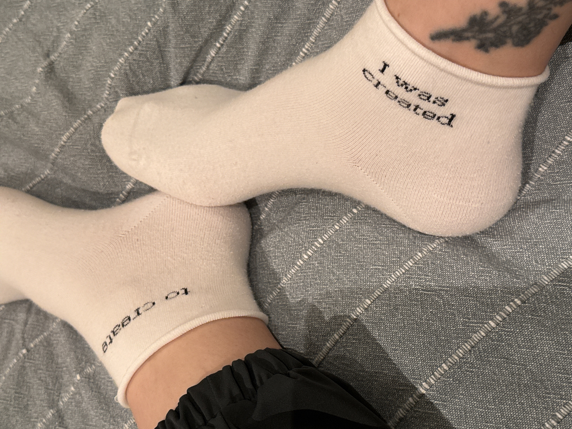 The image shows a close-up view of two feet wearing white ankle socks with black writing. The text on one sock reads "I was created" and the text on the other reads "to create." There is a tattoo visible on the right ankle. The feet are resting on a gray textured surface, likely a bedspread or blanket. The background is out of focus. There is no evidence in the image to suggest it was taken in Barcelona, Spain.