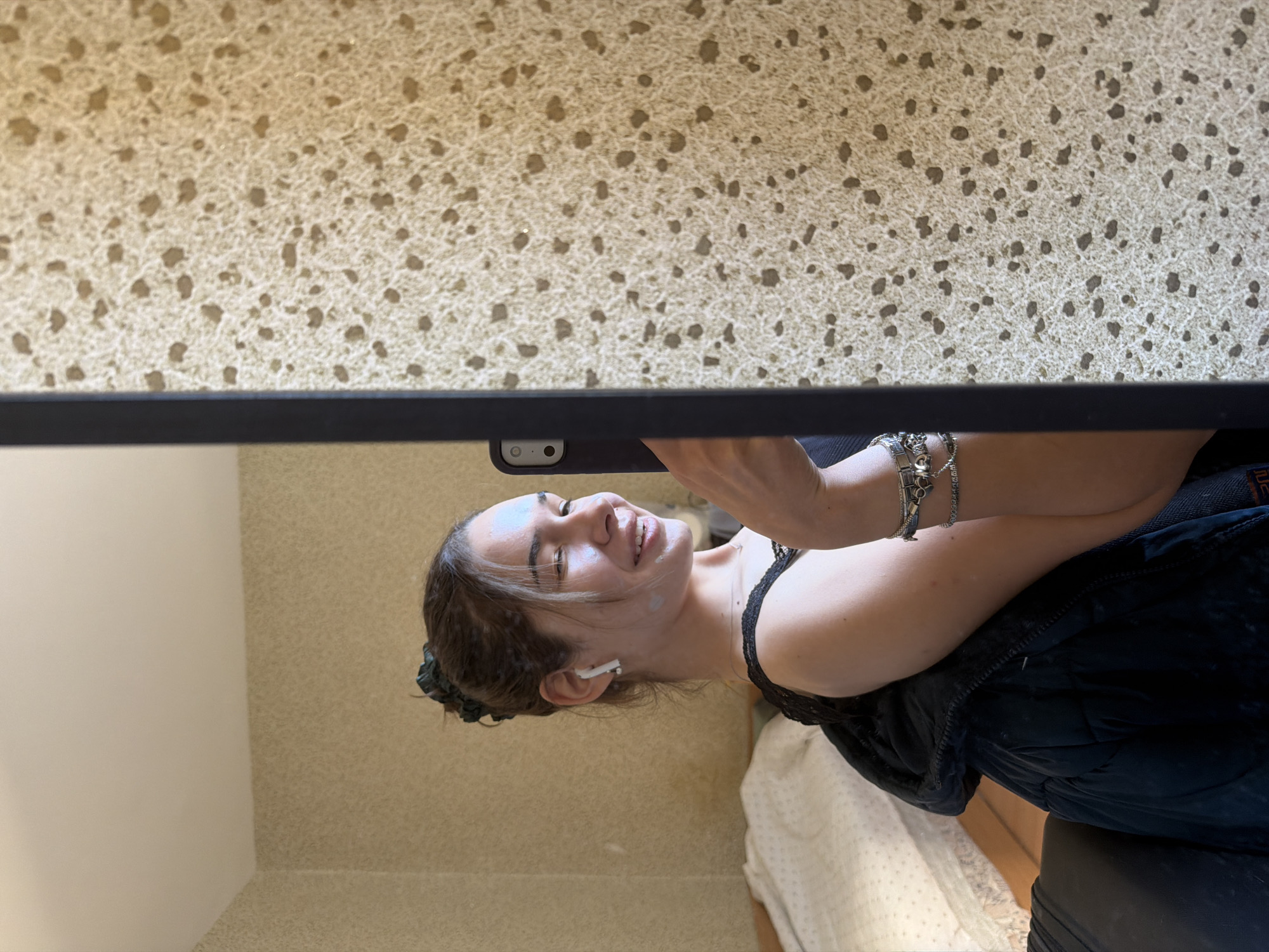 A young woman is captured in what appears to be a selfie, reflected in a surface above her. The image is oriented sideways, but if rotated, she is looking upwards and smiling. She has dark hair styled loosely, with some strands framing her face, and is wearing a single white earbud in her right ear. Her left arm is visible, adorned with several metallic bracelets on her wrist. She is wearing a black top, possibly a camisole or tank top, and a dark, possibly padded, jacket or coat. Her expression is joyful, with a bright smile.
The scene appears to be indoors during the daytime. The reflective surface above her has a textured, speckled pattern in light beige with irregular dark brown spots. A dark horizontal bar, possibly an edge or frame, runs across the mid-ground. Behind the woman, a plain, light-colored wall is visible, along with a glimpse of white fabric with a subtle pattern, perhaps bedding. No text is visible in the image. The specified location context is Sofia, Bulgaria, though this is not visually evident in the image itself.
