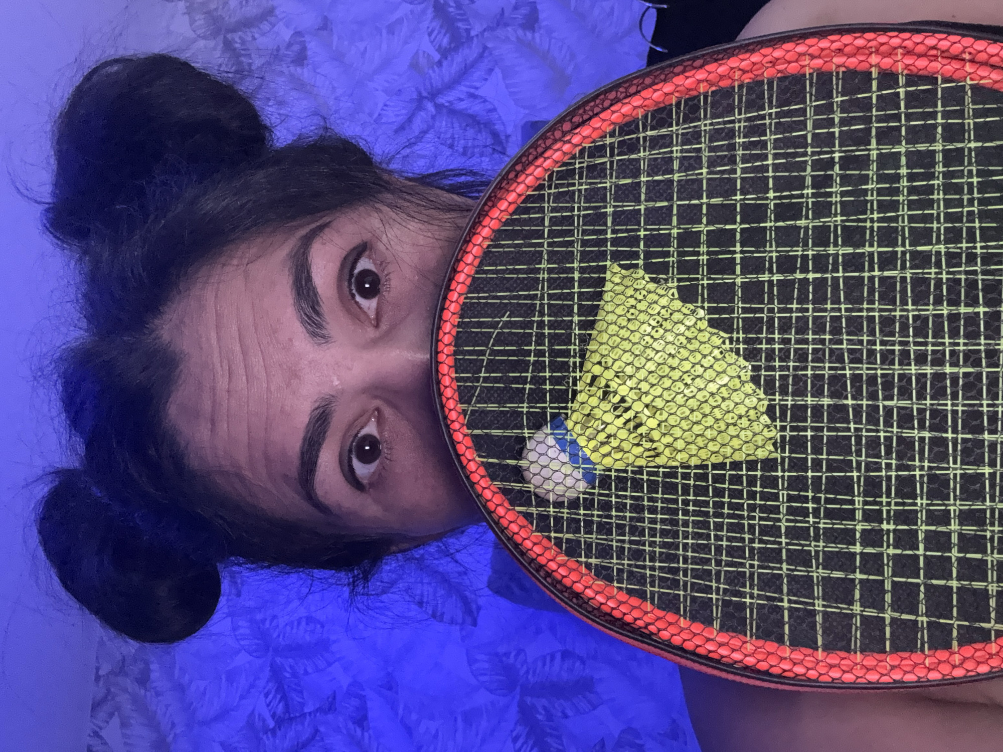 A person is visible in a close-up, top-down view, partially obscured by a badminton racket. The subject's face is positioned on the left side of the frame, with only the forehead and eyes visible as the badminton racket covers the lower half. The person has dark hair styled in two buns, one of which is clearly seen on the top left. Their eyes are wide open and directly looking at the viewer.
A black and red badminton racket occupies the right side of the image, its strings having a bright yellow/green hue. A yellow shuttlecock is caught in the strings of the racket, and a smaller white and blue object, possibly another shuttlecock head, is also visible near it.
The background consists of a blue or purple patterned fabric, likely bedsheets or a blanket, suggesting an indoor setting such as a bedroom. The entire scene is bathed in a strong blue ambient light, giving the image a distinctive cool tone. There is no visible text. The person appears to be posing for the camera, holding the racket with the shuttlecock integrated into the shot.