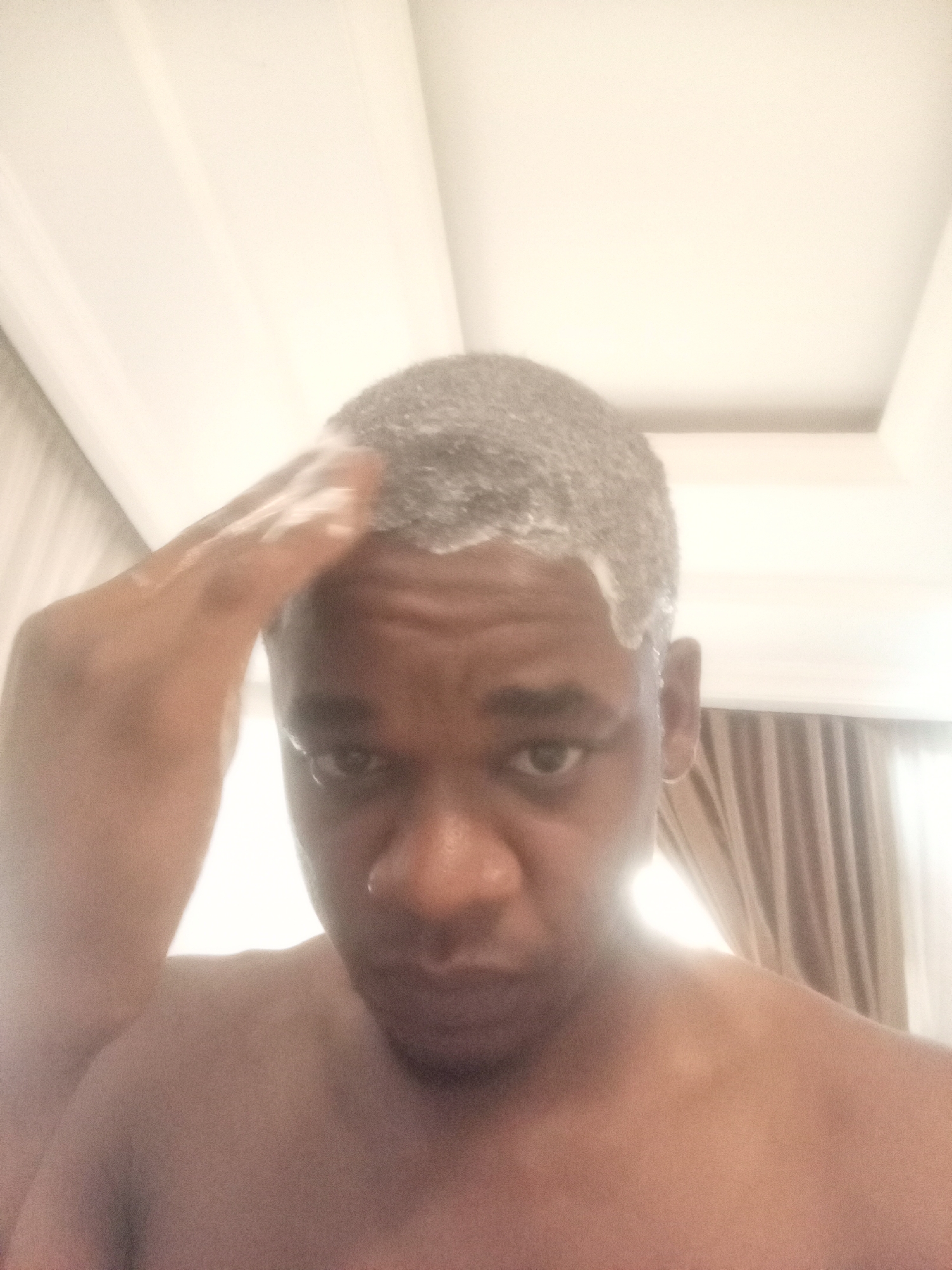 A close-up, slightly low-angle photograph depicts a Black man washing his hair. His hair is covered in a white, foamy substance, presumably shampoo or soap. He is touching his hair with his right hand, also covered in the white substance. The man appears to be indoors, with a white ceiling visible above him, featuring a recessed, rectangular design. A tan-colored curtain is visible in the background. The photograph is taken from a point-of-view perspective. The photograph does not include any evidence to suggest a specific location in Kubwa, Nigeria.