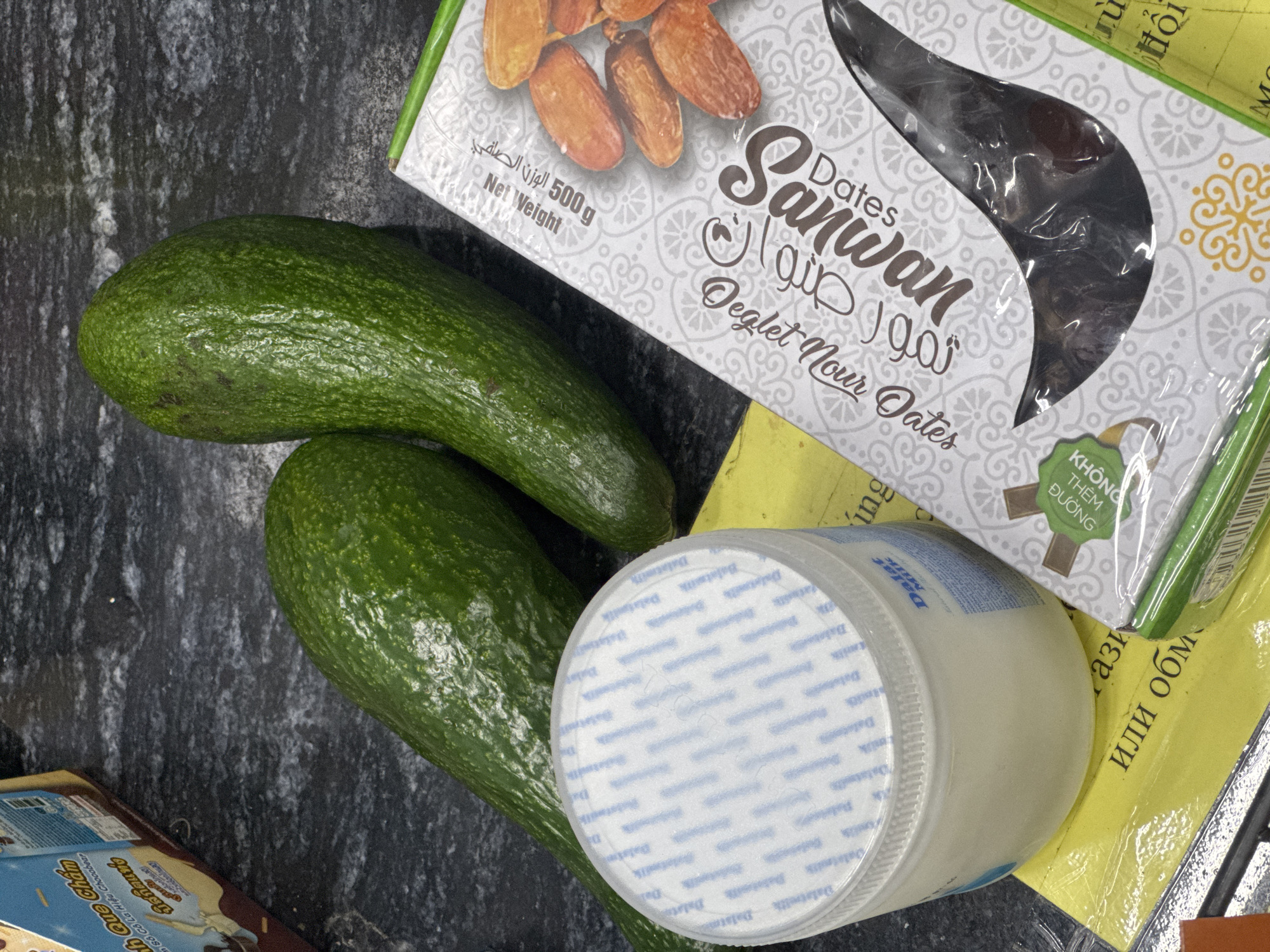 Two avocados are positioned on a dark, speckled countertop. To the right of the avocados, a package of dates with the brand name "Sanwan" is visible. In the foreground, a white plastic container with a blue and white patterned lid is situated on a yellow surface. The lid displays repeating text, possibly a brand name, in blue. Partially obscured on the yellow surface are some Russian Cyrillic characters. A corner of a brown and beige cardboard box with Vietnamese text is visible in the upper left of the frame. The overall scene suggests a kitchen or food preparation area. The provided information does not contain any location data for Bac Nha Trang Ward, Vietnam.