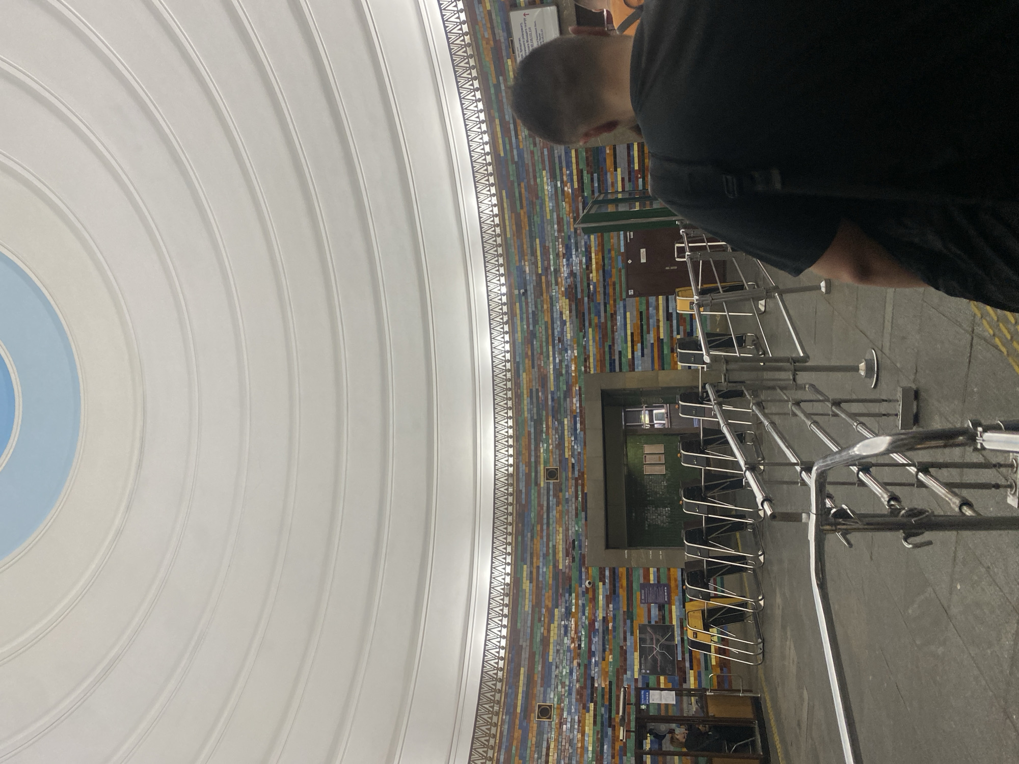 The image depicts the interior of a metro station, likely in an urban environment. The ceiling is a prominent feature, characterized by a series of concentric white rings that create a dome-like effect. At the apex of the dome, a section of light blue sky is visible, possibly an artistic representation or a skylight. The walls of the station are adorned with a vibrant mosaic of rectangular tiles in a variety of colors, including shades of blue, green, yellow, red, and brown, arranged horizontally in staggered rows. Above the tiled wall, a decorative frieze runs along the circumference.
In the foreground, a person wearing a black t-shirt and carrying a backpack is standing, facing away from the camera and towards the turnstiles. The turnstiles themselves are made of polished metal and are set up in a row, indicating an entry or exit point. To the left of the turnstiles, there is a booth with a person inside, and a display board with what appears to be a map. The floor of the station is paved with dark tiles. The overall impression is one of a public transportation hub with an emphasis on decorative architectural elements.