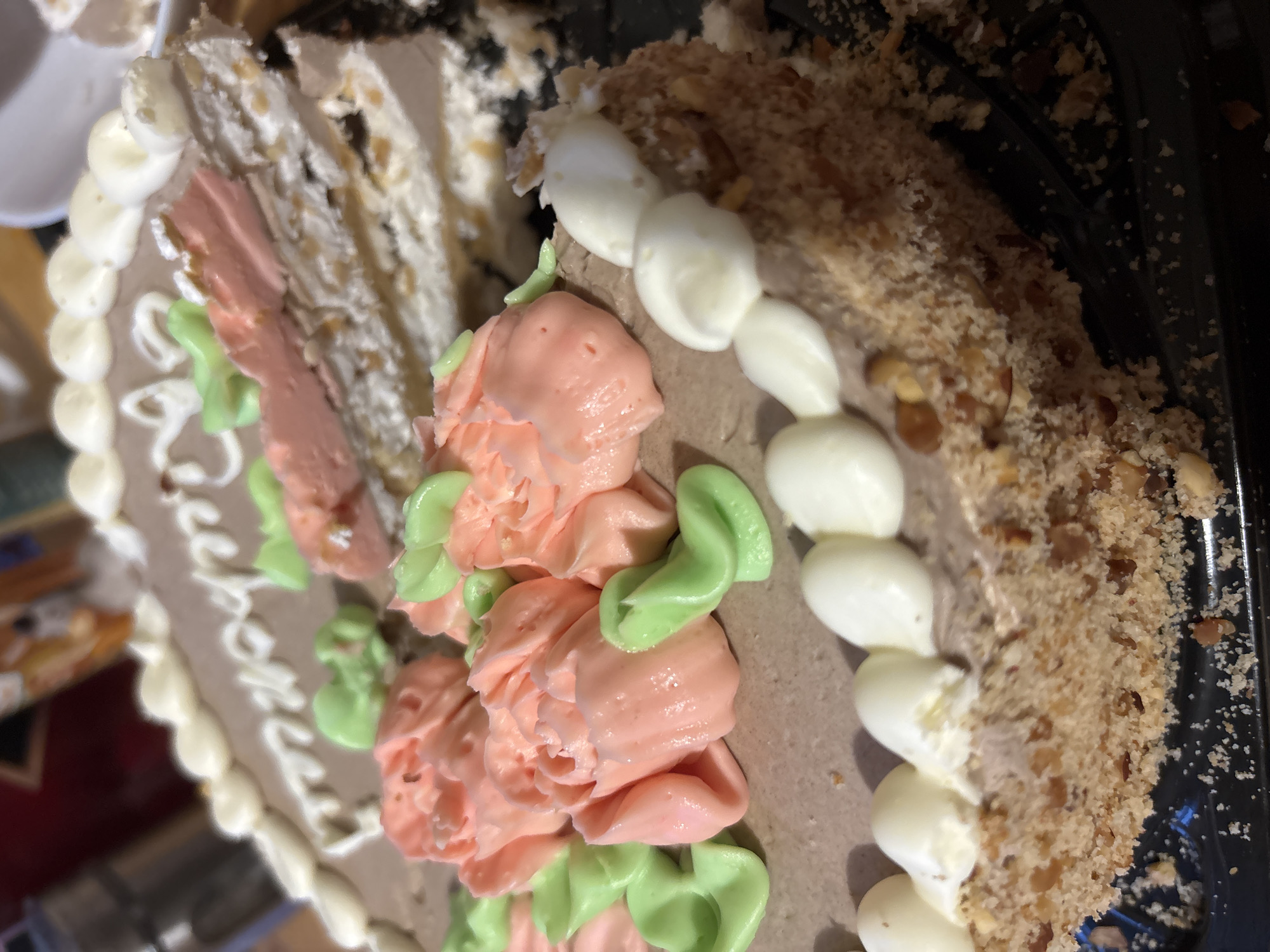 The image shows a slice of cake decorated with pink and green frosting flowers and a row of white frosting dollops along the edge. The sides of the cake are covered in a crumb-like texture with visible nuts. The cake is on a dark-colored serving tray. Part of the cake appears to have been cut and removed. The inscription "Happy Birthday" is written on the cake surface. The location is unknown.
