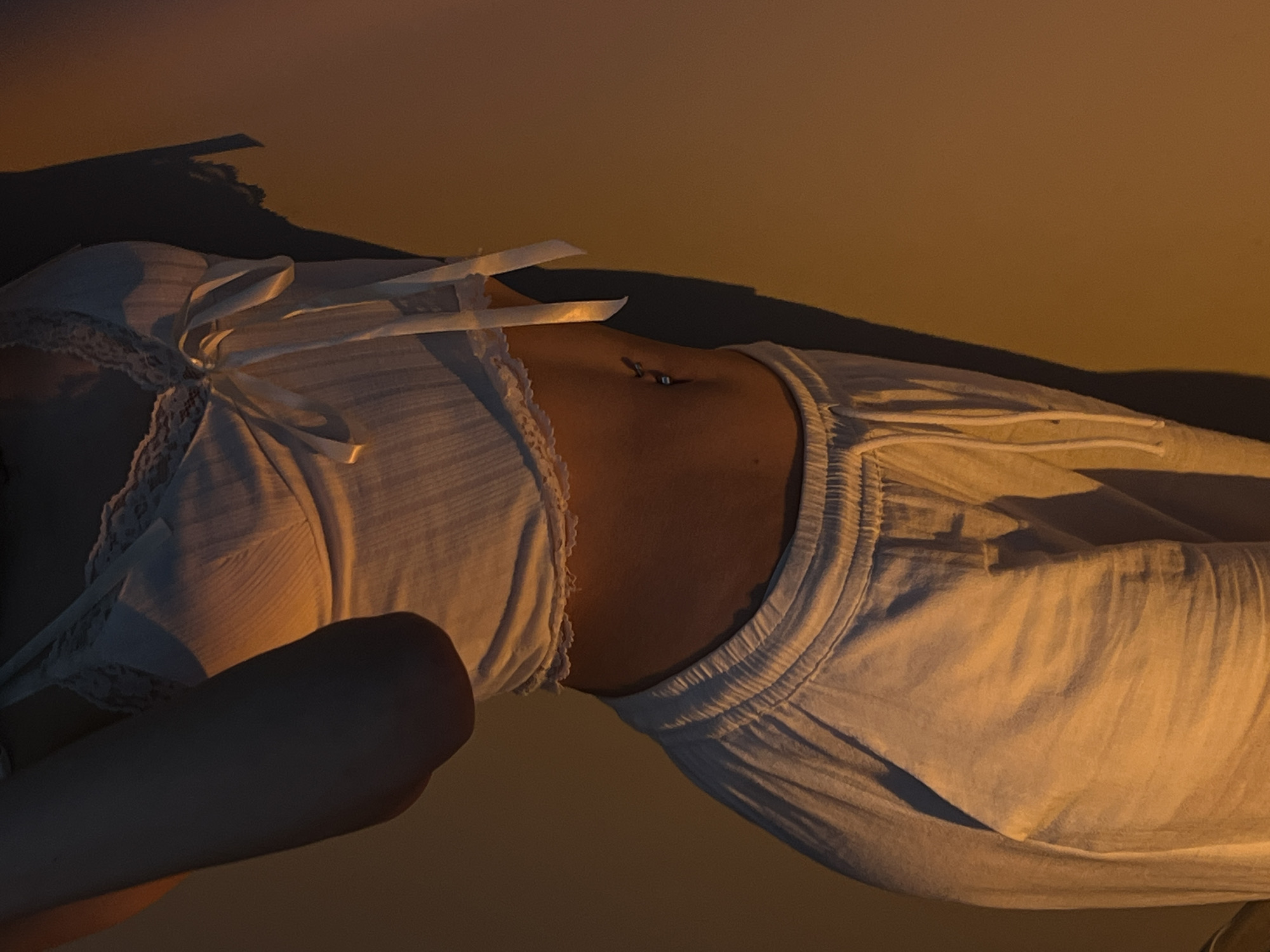 The image shows a close-up view of a person's torso, from the chest down to the thighs. The person is wearing a white lace-trimmed camisole with a bow at the front and white loose-fitting pants. The lighting is warm and dramatic, casting shadows on the wall behind the person. A navel piercing is visible. The scene does not provide any information about the city of Ternopil, Ukraine, or any specific location within it.