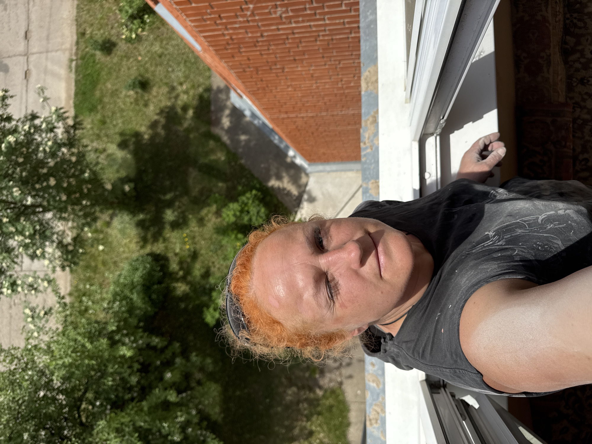 A photograph shows a fair-skinned person, likely a woman, positioned at a high angle looking down from a building or structure. The person has short, curly reddish hair, and is wearing a dark-colored t-shirt. Her skin appears dusty or lightly marked, possibly from work. The person is near a white window frame. Beyond the window, there is a view of green foliage, trees, and a sidewalk or pavement, suggesting an urban or suburban setting. The presence of a brick building immediately adjacent indicates an apartment or residential complex. Given the prompt specifying Chaikovsky, Russia, it's reasonable to assume the location is somewhere within this city, near a residential area with buildings of similar construction.