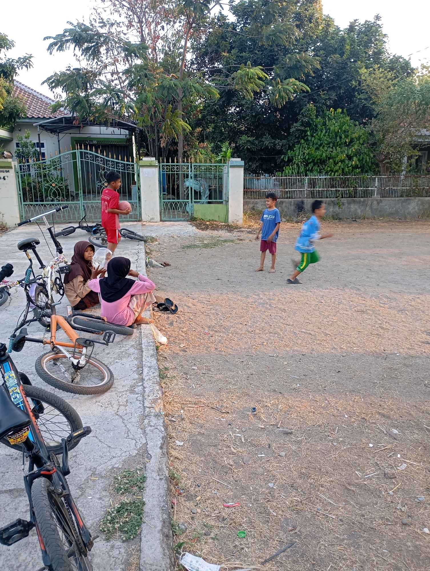 In Babelan, Indonesia, on a sun-drenched, dusty street, a group of children are engaged in outdoor activities. In the foreground, several bicycles are parked or resting on a concrete curb, some in disarray. Three young children are seated on the curb. One, wearing a pink hijab and a pink long-sleeved shirt, is looking to the side, holding a large transparent plastic bottle. Another child, in a brown hijab and a brown uniform-like shirt, is also seated. A third child, wearing a red shirt with the number '10' and red shorts, stands holding a small reddish ball.
Further down the street, two other boys are active. One, in a blue t-shirt and red shorts, stands barefoot, looking towards the other boy. This other boy, blurred due to motion, is running with his arms extended, wearing a blue t-shirt and green shorts with yellow stripes.
The background features a residential house with a tiled roof, a green metal fence with decorative elements, and lush green trees and foliage. The ground is a mix of dry earth, scattered straw, and some debris. The scene captures a moment of childhood play in a suburban environment.