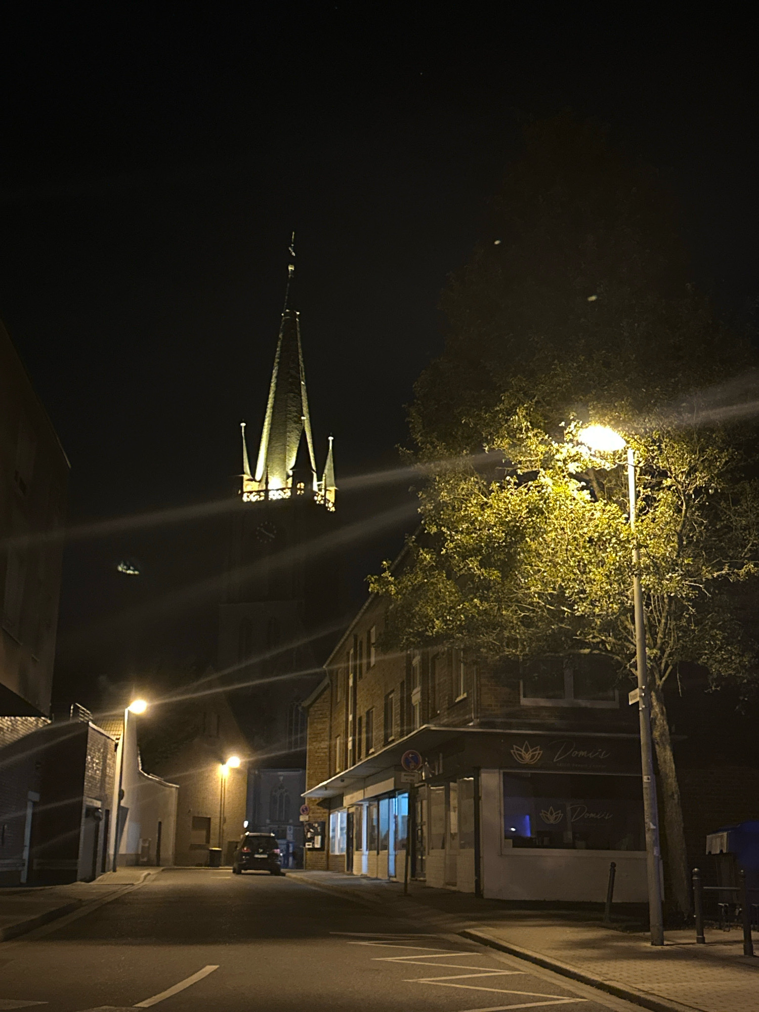 The image shows a street scene at night in Krefeld, Germany. A church spire with illuminated details is visible in the background, towering over the surrounding buildings. A street lamp with a bright light illuminates a tree with dense foliage on the right side of the image. On the left, buildings line the street, with shop windows displaying illuminated signage that reads "Domis". A dark-colored car is parked on the street. The street is paved, and there are pedestrian sidewalks. The sky is dark, indicating nighttime.
