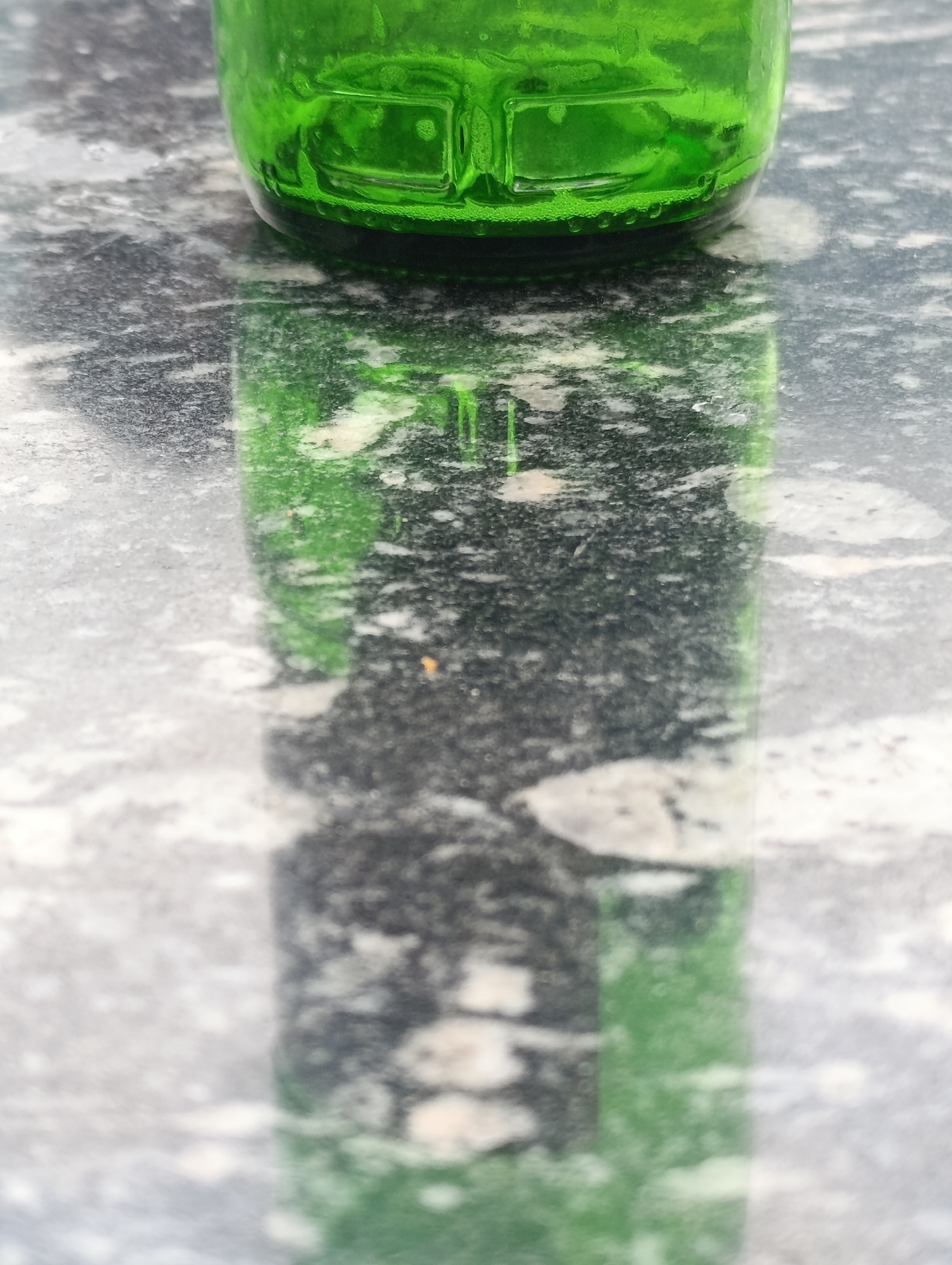The image shows the bottom portion of a green glass bottle sitting on a speckled, dark surface, likely a marble tabletop. The bottle is reflecting onto the surface below, creating a distorted, elongated green shape. The surface has a mottled pattern of grey, black, and white. The upper part of the bottle shows some liquid and small bubbles. The lighting appears to be natural, casting shadows and highlights on the bottle and its reflection. There are no people or any indications of a specific city or location like Old Manali, India present in the image.