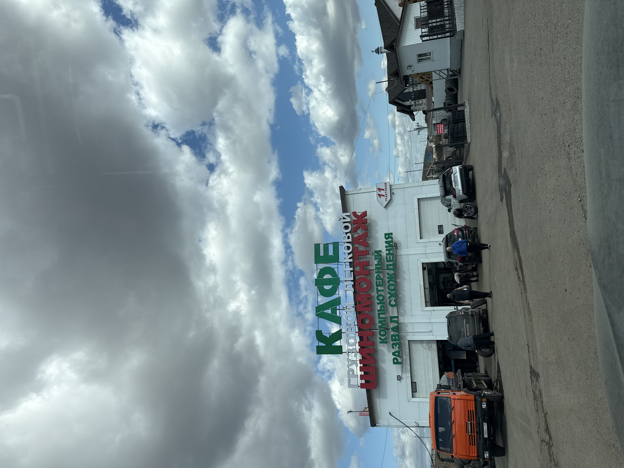 The image shows a tire shop and cafe in Дрокино, Russia. The sign on the building reads "КАФЕ ГРУЗОВОЙ ШИНОМОНТАЖ" (Cafe Truck Tire Service) and "КОМПЬЮТЕРНЫЙ РАЗВАЛ-СХОЖДЕНИЯ" (Computer Wheel Alignment). Several people are working on vehicles parked in front of the building. There is an orange Kamaz truck, a black SUV, a silver car, and other vehicles. The building is white with multiple garage doors. The sky is cloudy with patches of blue. A building with a dark roof is located next to the tire shop.
