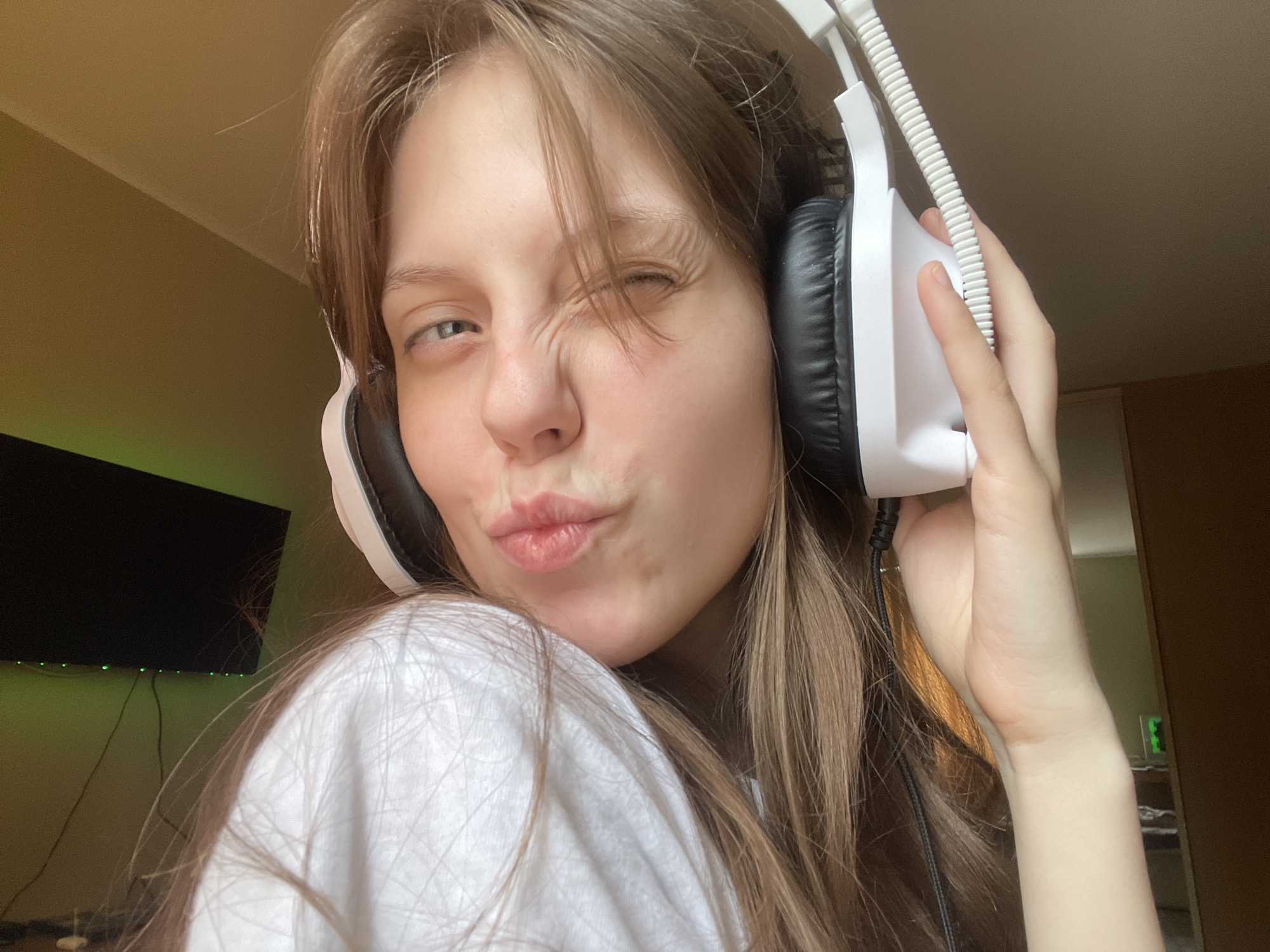 A young woman, with long brown hair and fair skin, is captured in a close-up selfie. She is wearing a white t-shirt and large white and black headphones. One eye is closed in a wink, while the other is open, looking towards the camera. Her lips are pursed in a kissing gesture. Her left hand is visible, holding the headphones near her ear. In the background, a black television screen is visible on the left, with subtle green lighting. The wall behind her appears to be a warm, muted yellow. The overall setting suggests an indoor environment, possibly a room in an apartment. There are no discernible landmarks or indicators that place the image specifically in Ufa, Russia.