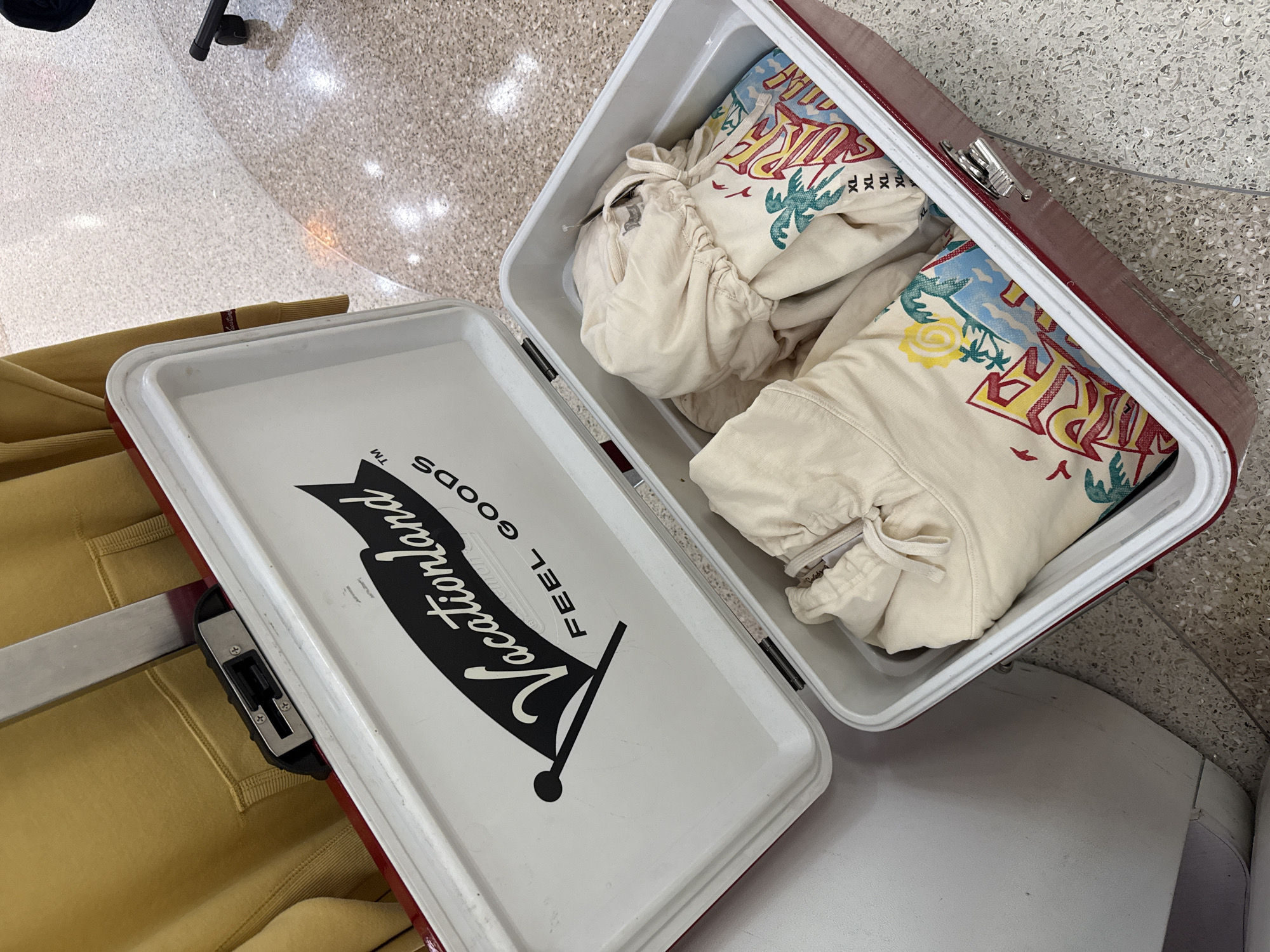 A high-angle shot depicts an open maroon and white suitcase with contents, labeled "Vacationland," with a yellow garment laying to the left of the suitcase. The suitcase is filled with what appears to be bundled clothing, bearing a "Surf" design with palm trees. The surroundings consist of tiled flooring and the base of a chair is visible in the background. This image was taken in Fort Lauderdale, United States of America.