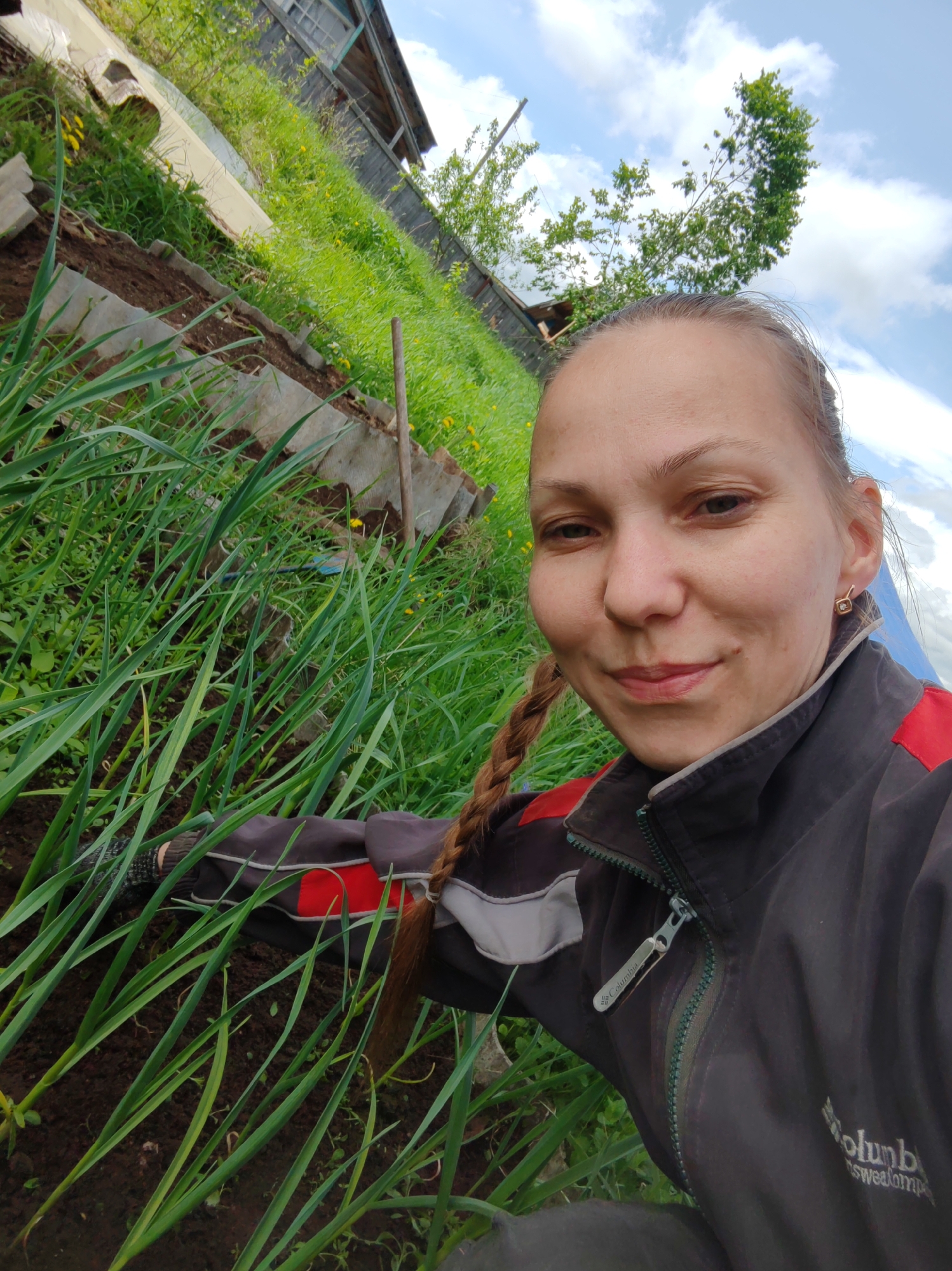 A selfie shows a woman in Чухлэм, Russia, in what appears to be a garden. She has a braid over her shoulder and is wearing a black and red jacket. She is smiling slightly. A row of tall green plants, likely onions or garlic, grows in the foreground. In the background, there is a sloped grassy area leading up to a wooden building. The sky is visible in the upper right, with some clouds.