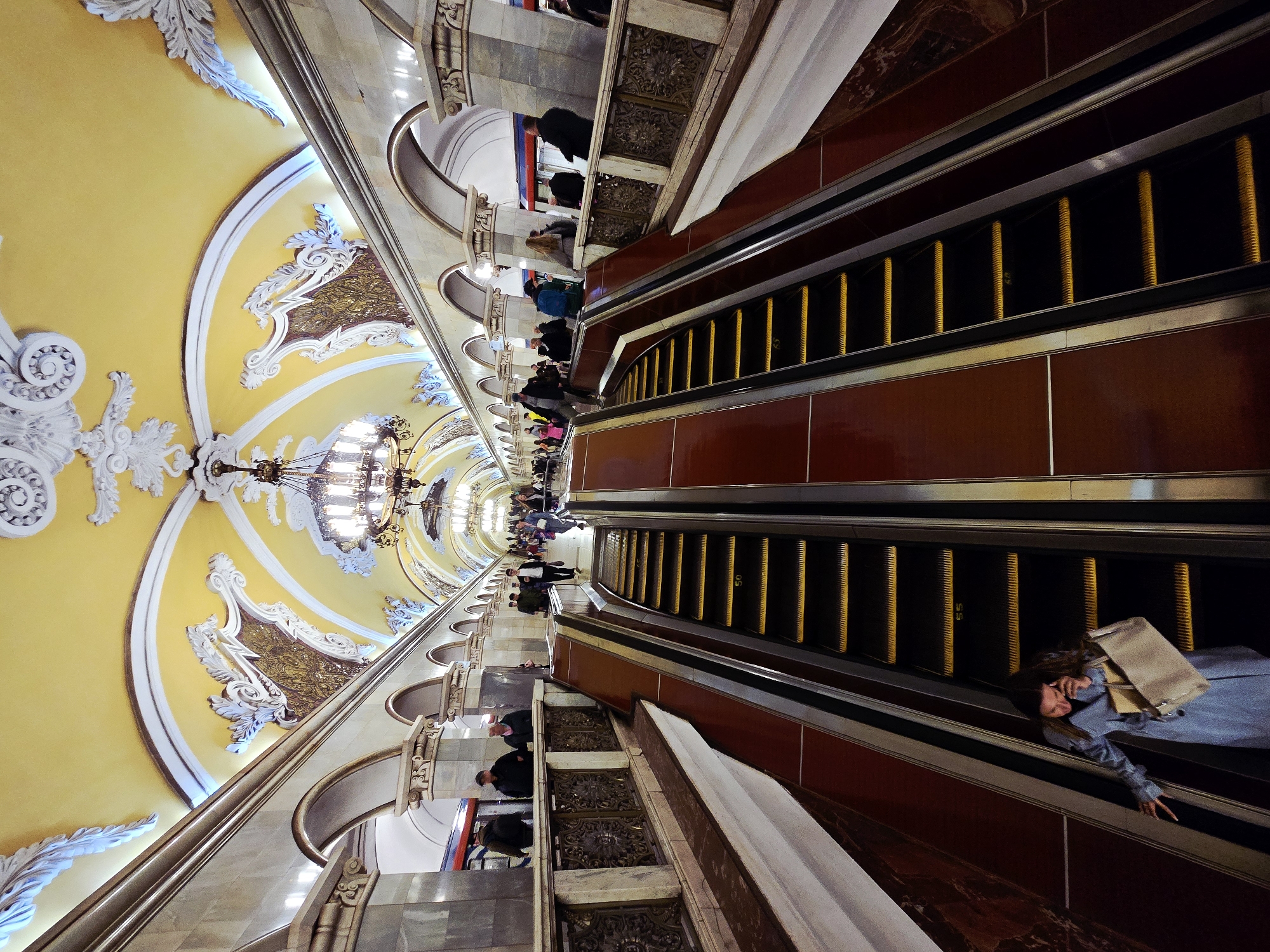 The image shows an interior view of a Moscow Metro station in Russia. The scene is shot from a low angle, looking upwards along an escalator leading into the station. The station features elaborate architectural details, including ornate chandeliers, a high vaulted ceiling painted yellow and decorated with white baroque-style stucco reliefs, and marble-clad walls. Two escalators run parallel, with people riding up and down. The escalator sides are clad with dark red ceramic tiles. A woman in a blue jacket and carrying a shopping bag is visible riding the escalator closer to the foreground. People can be seen standing and walking on the station platform in the distance.