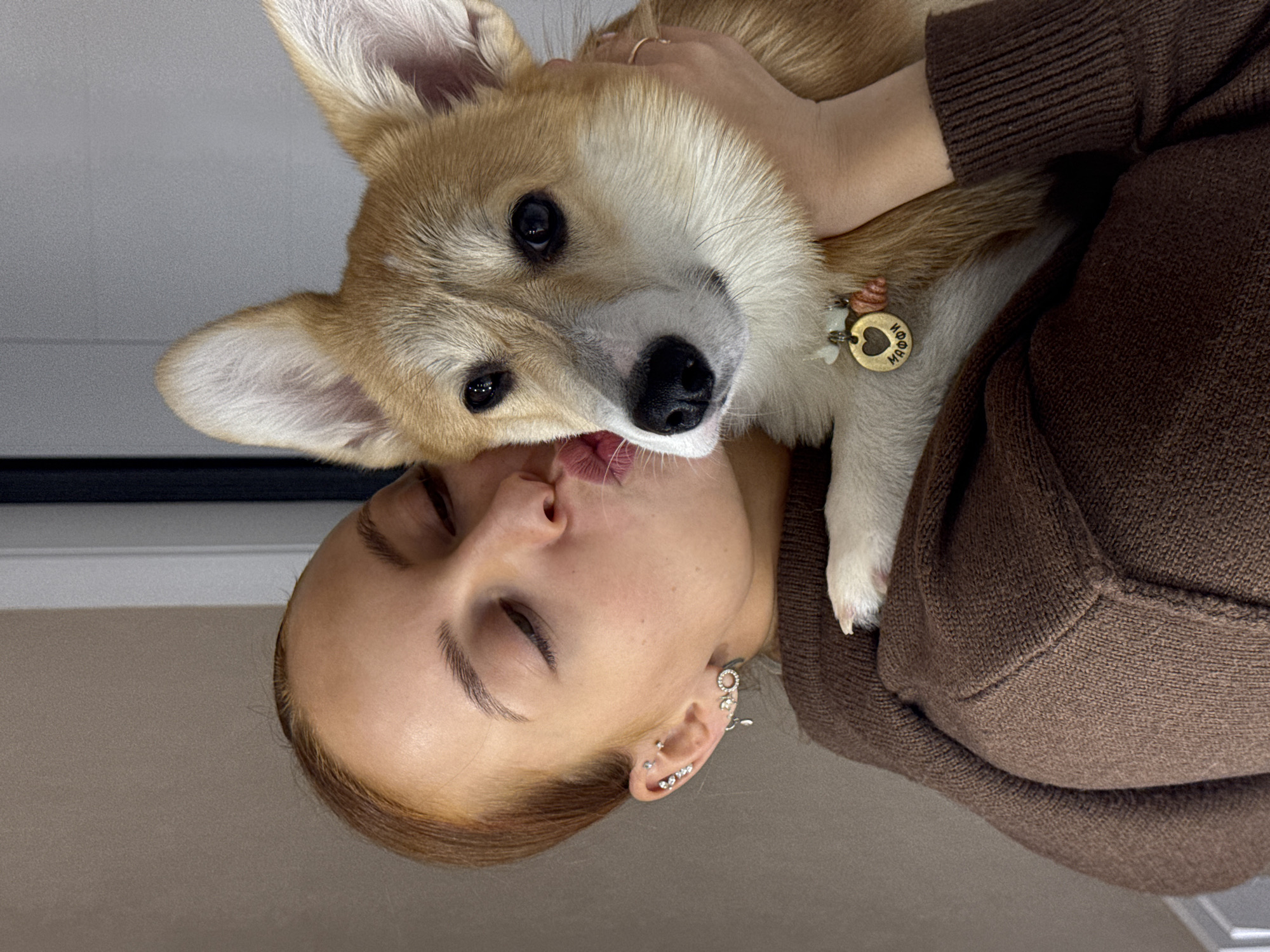 A heartwarming close-up portrait captures a moment of affection between a person and a dog, set indoors. The image depicts a young adult female embracing and kissing a small, light-brown and white dog, likely a Corgi.
The person, positioned with her head tilted, has light brown hair and fair skin. Her eyes are gently closed, and her lips are pursed as she presses them against the dog's snout. She is wearing a textured brown knitted sweater. Her left ear is visible, adorned with multiple silver piercings, including studs, a small hoop, and a decorative cuff.
The dog, held close, looks directly at the camera with large, dark eyes. Its fur is a mix of light brown, cream, and white, characteristic of a Corgi. Around its neck, a collar is visible, featuring a heart-shaped metal tag and a small, brown, curled charm. The heart-shaped tag has visible Cyrillic text that reads "МОЙ МАЛ" (Moy Mal), which translates to "My Little One" or "My Boy."
The background is simple, consisting of a light gray or off-white wall in the upper half of the frame, transitioning to a darker, indistinct surface at the bottom. The lighting is soft and even, suggesting an intimate indoor setting. The scene conveys a strong sense of warmth, love, and contentment between the owner and their pet, captured in what appears to be a home environment in Chaikovsky, Russia.
