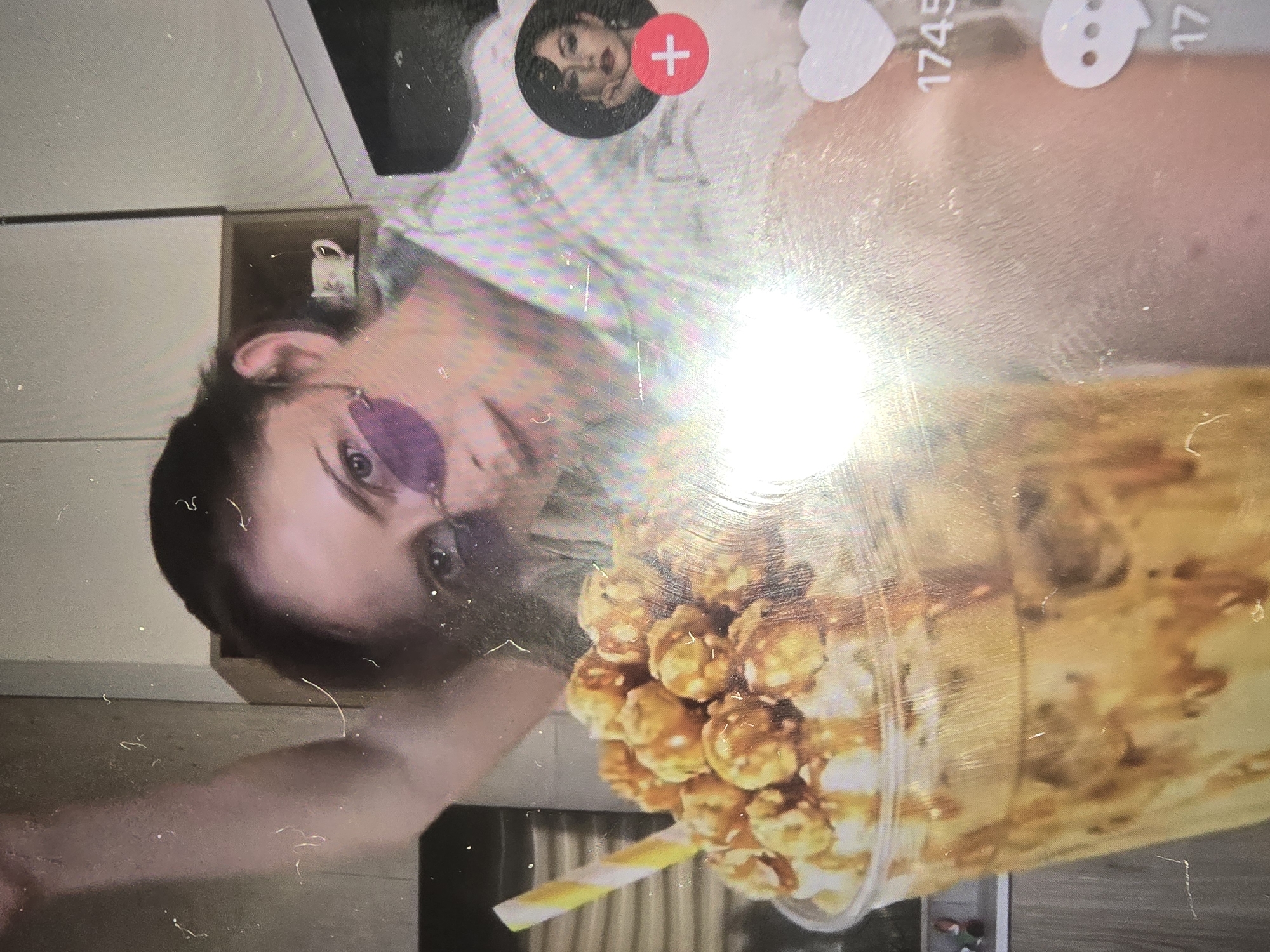 The image shows a close-up of a drink topped with caramel popcorn, held by a person wearing a white, patterned shirt and purple tinted glasses. The person's face is partially visible, with dark hair and light skin. In the background, a shelf with small decorative items can be seen. The overall scene appears to be indoors, possibly a room or an office. The image is also characterized by numerous scratches and dust particles on the surface, indicating it might be a photograph of a screen or a print. There is no identifiable information within the image to suggest a specific location in Ulan-Ude, Russia, or any other city.