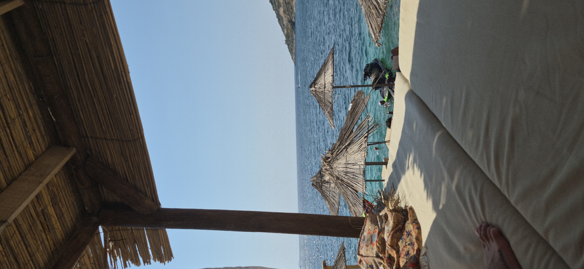 The image depicts a serene beach scene at a resort in Saranda, Albania. In the foreground, a person's bare foot rests on a padded lounge chair. The chair is positioned under a thatched roof structure, supported by wooden pillars, offering shade from the sun. Beyond the lounge chair, the turquoise waters of the Ionian Sea stretch towards the horizon, meeting a clear blue sky. Two thatched umbrellas stand partially submerged in the shallow water, casting shadows on the sandy seabed. A jet ski is anchored nearby, hinting at recreational activities available at the resort. In the distance, a hilly coastline with sparse vegetation is visible under the bright sunlight. The overall impression is one of a tranquil and idyllic beach resort setting.