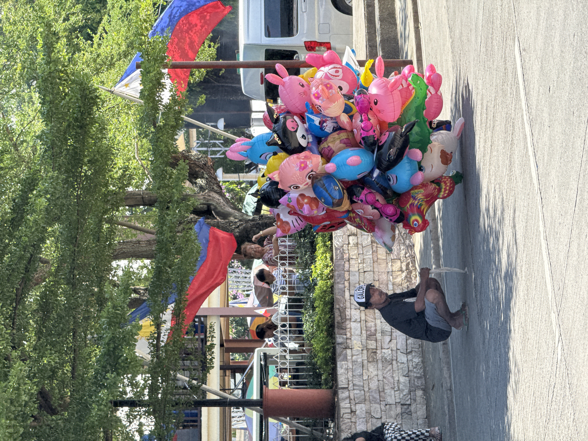 The image depicts a street scene in Davao City, Philippines, under bright sunlight. A man wearing a black t-shirt, gray shorts, and a black cap with the word "GALOP" is crouching on the pavement. He is holding a string attached to a large cluster of colorful inflatable balloons, many of which are shaped like cartoon animals such as rabbits, fish, and possibly a Hello Kitty character. The balloons are a mix of pink, blue, yellow, black, and red.
Behind the man, there is a low stone wall and a grassy area with bushes. Further back, a white van is parked on the right side of the frame. In the background, several people are visible near some structures, with umbrellas and Philippine flags draped in the background, suggesting a public area or an event. Trees with dense green foliage provide shade over parts of the scene. The ground is paved with asphalt and concrete, with shadows cast by the trees and possibly other unseen objects.