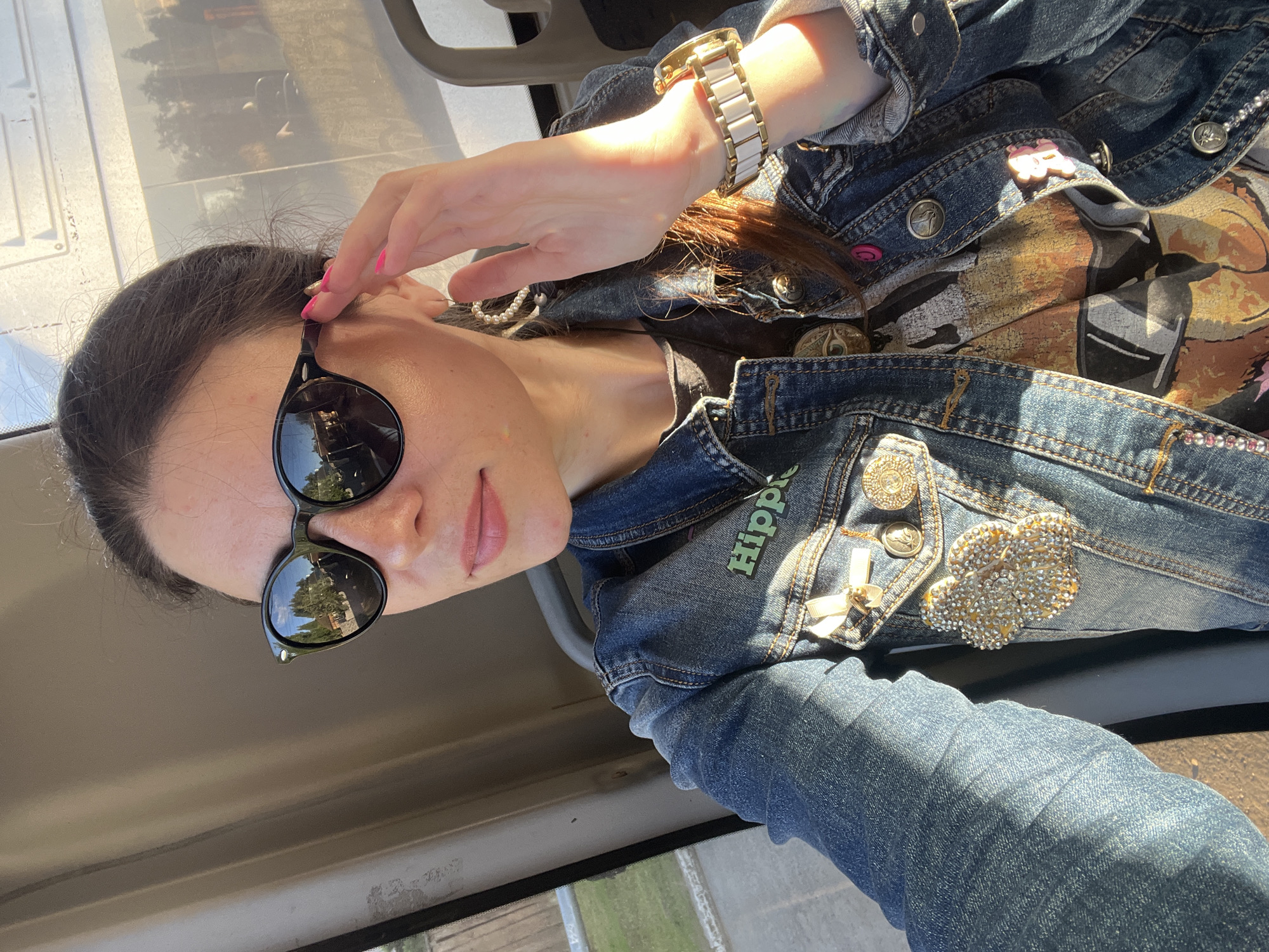This is a medium shot, rotated image of a young woman with dark hair, wearing sunglasses and a denim jacket. She is indoors, likely in a vehicle, as indicated by the window and the reflections in her sunglasses showing trees and a bright sky, suggesting it's daytime. The woman is touching her hair, looking towards the camera. Her denim jacket is adorned with several pins, including a "Hippie" pin, a pink smiley face pin, and a bejeweled flower pin. She is also wearing a gold and white watch on her left wrist. The overall mood is casual and sunny. The reflection in the sunglasses provides a glimpse of the outside environment.