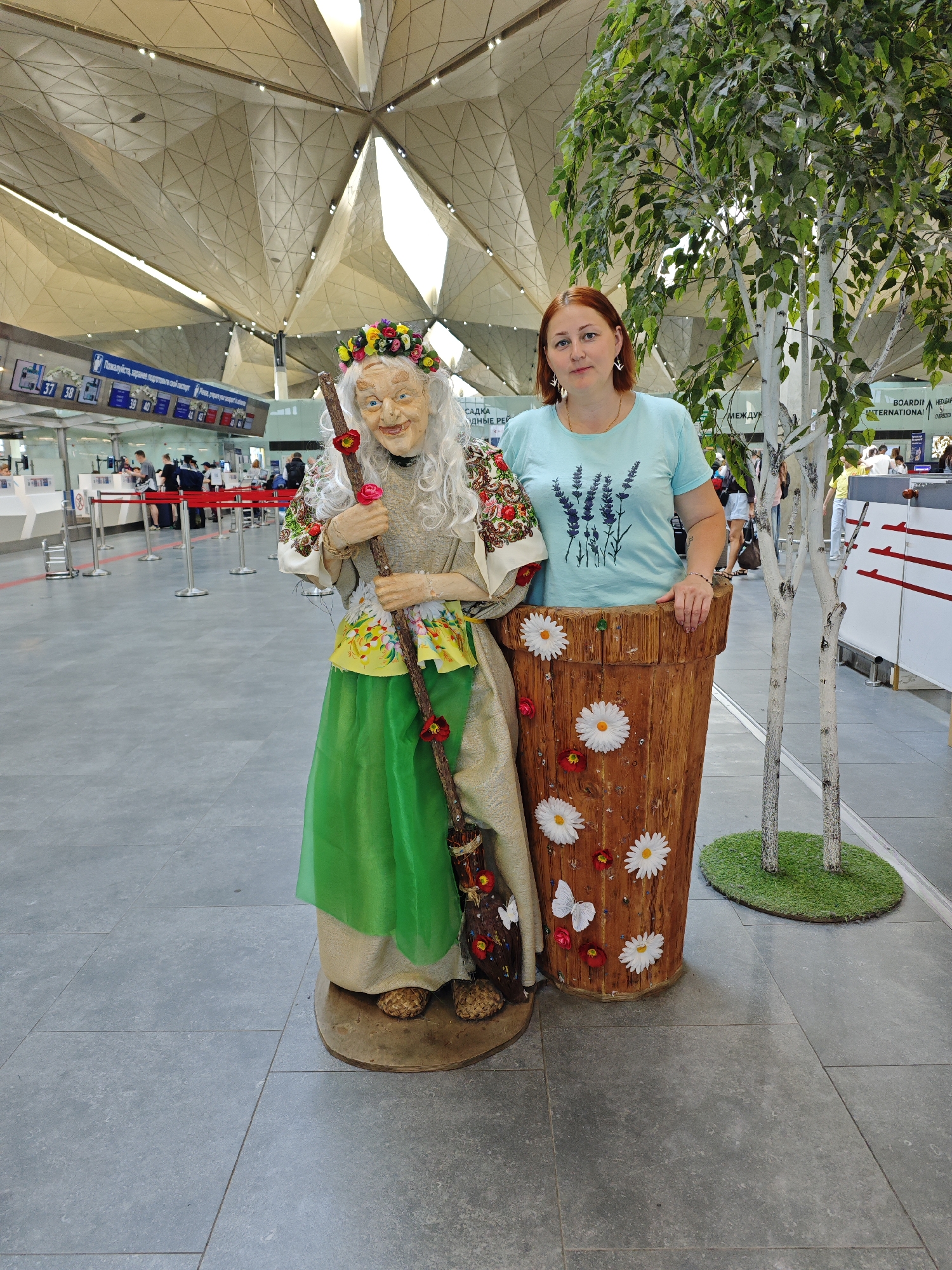 A woman with red hair stands next to a life-sized doll of Baba Yaga, the Russian folklore witch, in a modern indoor space with a distinctive geometric ceiling structure. The doll is dressed in traditional Russian attire, including a floral wreath on its head, a patterned headscarf, and a green skirt. It holds a broomstick adorned with flowers. The woman wears a light blue t-shirt with a lavender design and leans against a large, wooden planter decorated with artificial daisies and butterflies. Two slender, white-barked artificial birch trees are positioned behind the planter, with a patch of artificial grass at their base. In the background, airport-style check-in counters and signage are visible, suggesting the location is Pulkovo Airport in Saint Petersburg, Russia. People can be seen in the distance near the counters. The floor is tiled in a grey stone pattern.