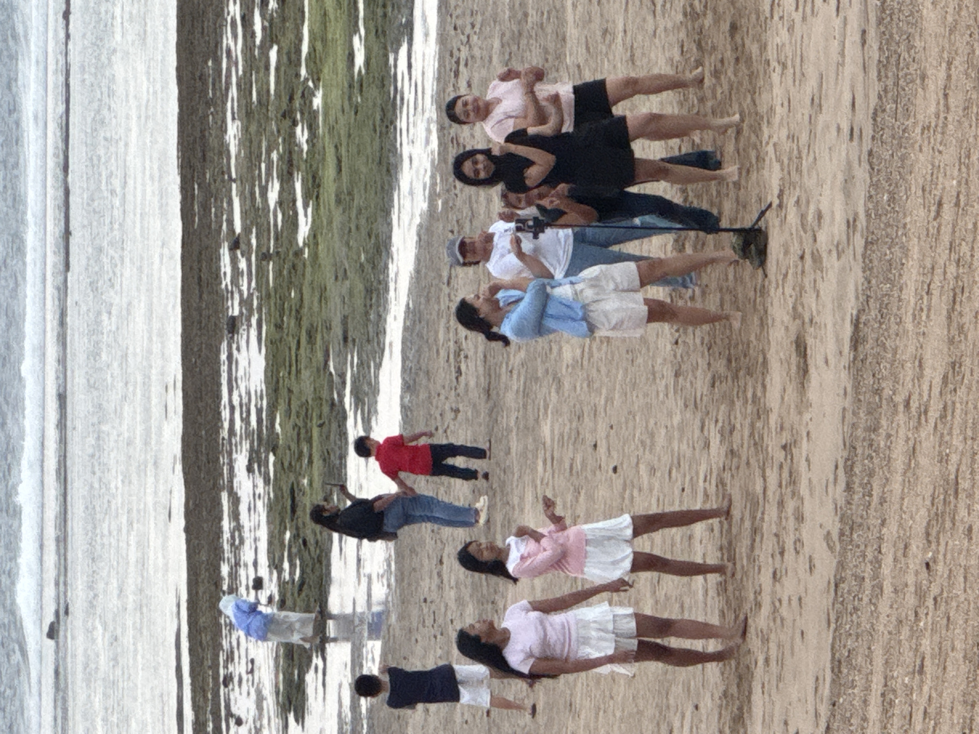 The image depicts a group of people on a sandy beach in Kuta Selatan, Indonesia, with the ocean and rocky intertidal zones in the background. In the foreground, four young women are gathered on the sand, with two standing closer and appearing to pose or interact. Behind them, a woman in a blue top and white shorts gestures while a woman in a white shirt and jeans stands next to her. Further back, a woman in a black dress stands with her arms crossed. To the left, two young girls in white skirts are standing, and a young boy in a blue shirt is walking away. In the middle ground, a woman in a dark top and jeans holds a phone, and a young boy in a red shirt holds her hand. In the far background, a person in a blue garment is visible near the water's edge, interacting with the environment. The scene appears to be a casual gathering, possibly a family outing or a group of friends, enjoying a day at the beach.