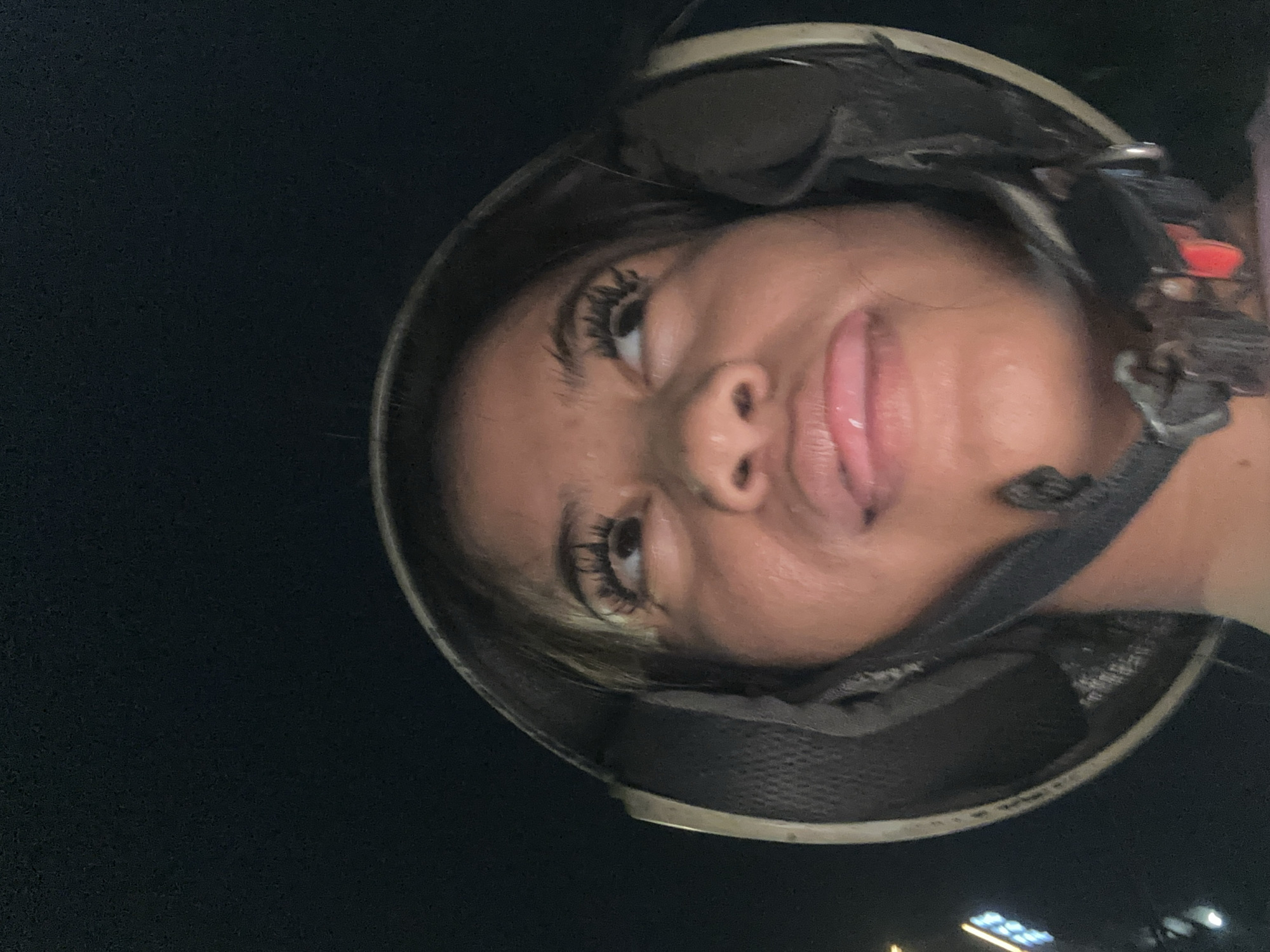 A woman is wearing a helmet and sticking her tongue out. The background is dark. There is some light in the lower right corner of the image. Based on the lighting conditions, it appears that the photo was taken at night. The location in the city of Central Jakarta, Indonesia is not discernible from the image.