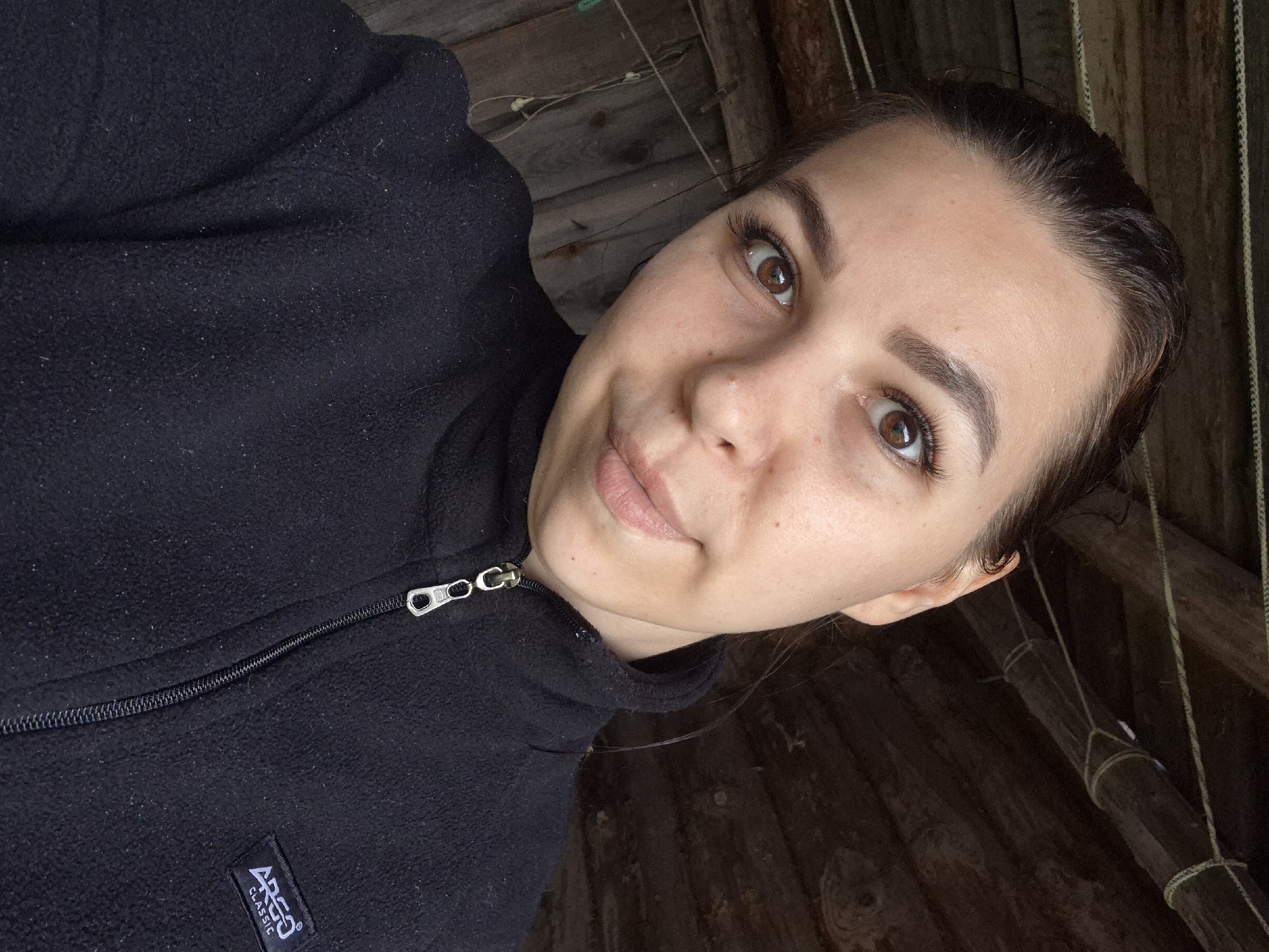 The scene is located in the city of Kharnavy, Russia. The image presents a close-up selfie of a young adult female with dark hair pulled back, brown eyes, and a slight smirk or pout. Her skin shows minor blemishes and small moles. She has well-defined eyebrows and long eyelashes. She is wearing a black, full-zip fleece jacket featuring a metallic zipper pull and a black rectangular patch with the white text 'ARGO CLASSIC' on the right chest area. The fleece fabric exhibits small white fibers and lint. The background is a rustic, dimly lit interior or sheltered outdoor space constructed from dark, weathered wooden planks and beams. Yellowish-white ropes are visibly strung and knotted around the overhead wooden beams. Thin wires or cords are also present against the wooden wall on the left side of the frame.