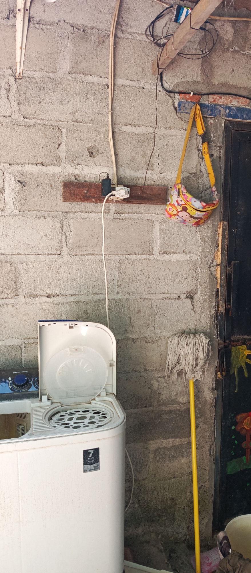 The image depicts an outdoor scene in Jatisari, Indonesia, focusing on a wall made of concrete blocks. Mounted on the wall is a wooden plank with an electrical outlet and a black adapter plugged into it, connected to a white electrical cord that runs downwards. To the right of the outlet, a small, yellow-strapped bag with a colorful pattern featuring cartoon characters is hanging from the wall. In the foreground, a white washing machine with its lid open is partially visible on the left. Next to the washing machine, a yellow mop with white strings stands upright. A dark metal door is visible on the right side of the image, with some colorful artwork or decorations on it. The ground appears to be concrete, with some debris and plastic items scattered around. The overall setting suggests a utility area or the exterior of a building.