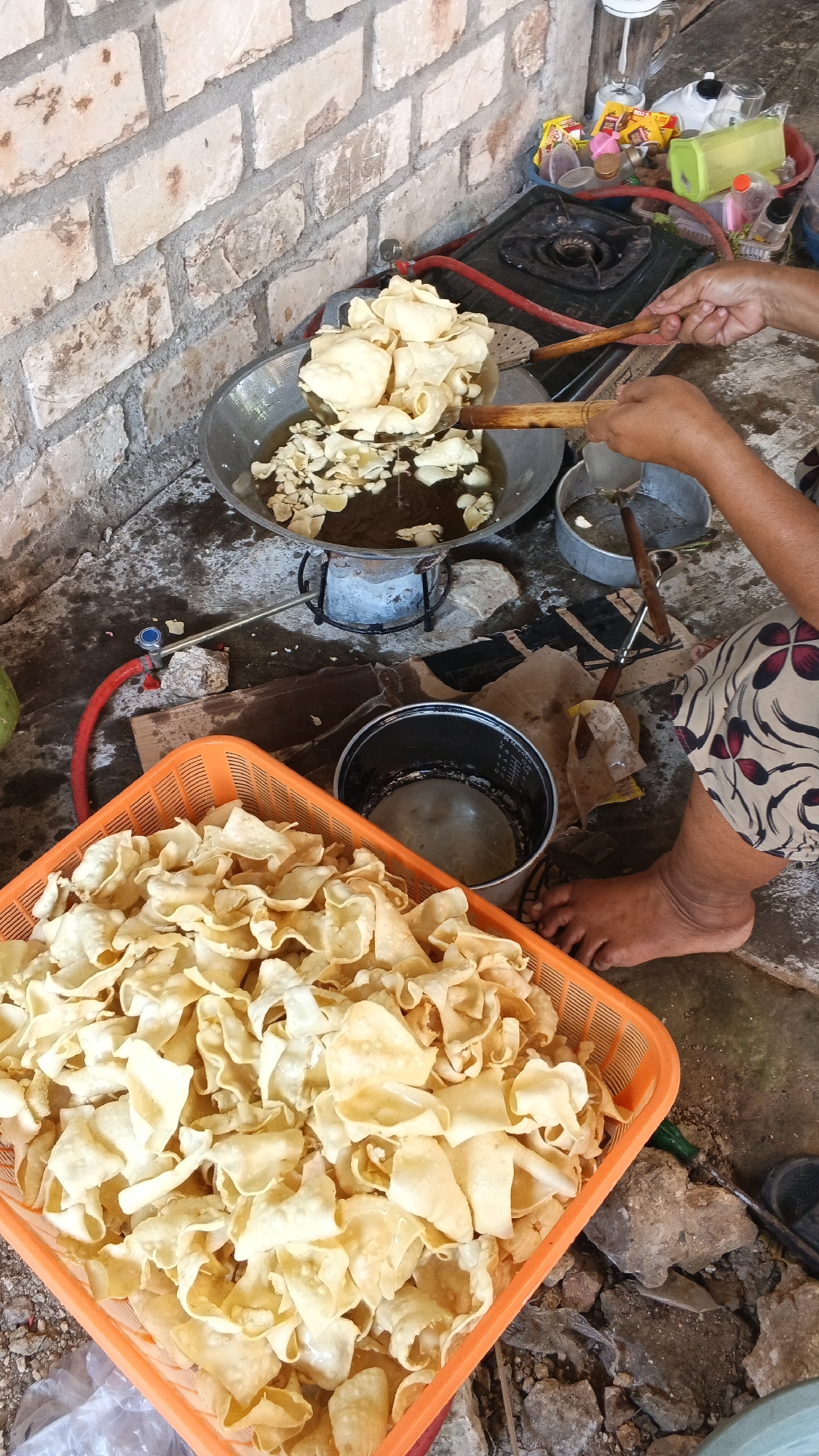 In Soko, Indonesia, a person is frying crackers in a wok over a gas stove. The wok is filled with oil, and the crackers are golden brown. A basket full of fried crackers is in the foreground, and a pile of raw crackers is in the wok. The scene is set outdoors, with a brick wall in the background. The ground is concrete, and there are a few rocks and debris scattered around. The person's feet are visible, and they are barefoot. The person is wearing a patterned skirt. A red gas hose is connected to the stove. There are several containers and bottles on a shelf behind the stove.