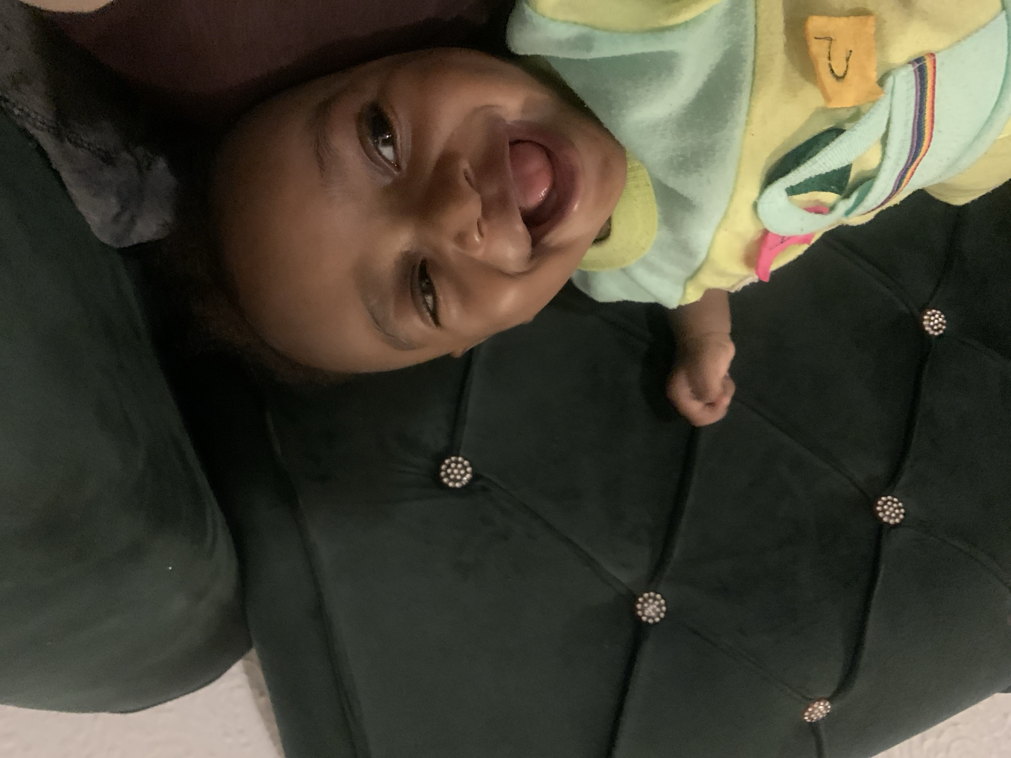 A baby lies on a dark green, tufted couch with decorative buttons. The baby is wearing a light yellow and green outfit with colorful accents. The baby is smiling with their mouth open, showing their tongue. The exact location is unknown.